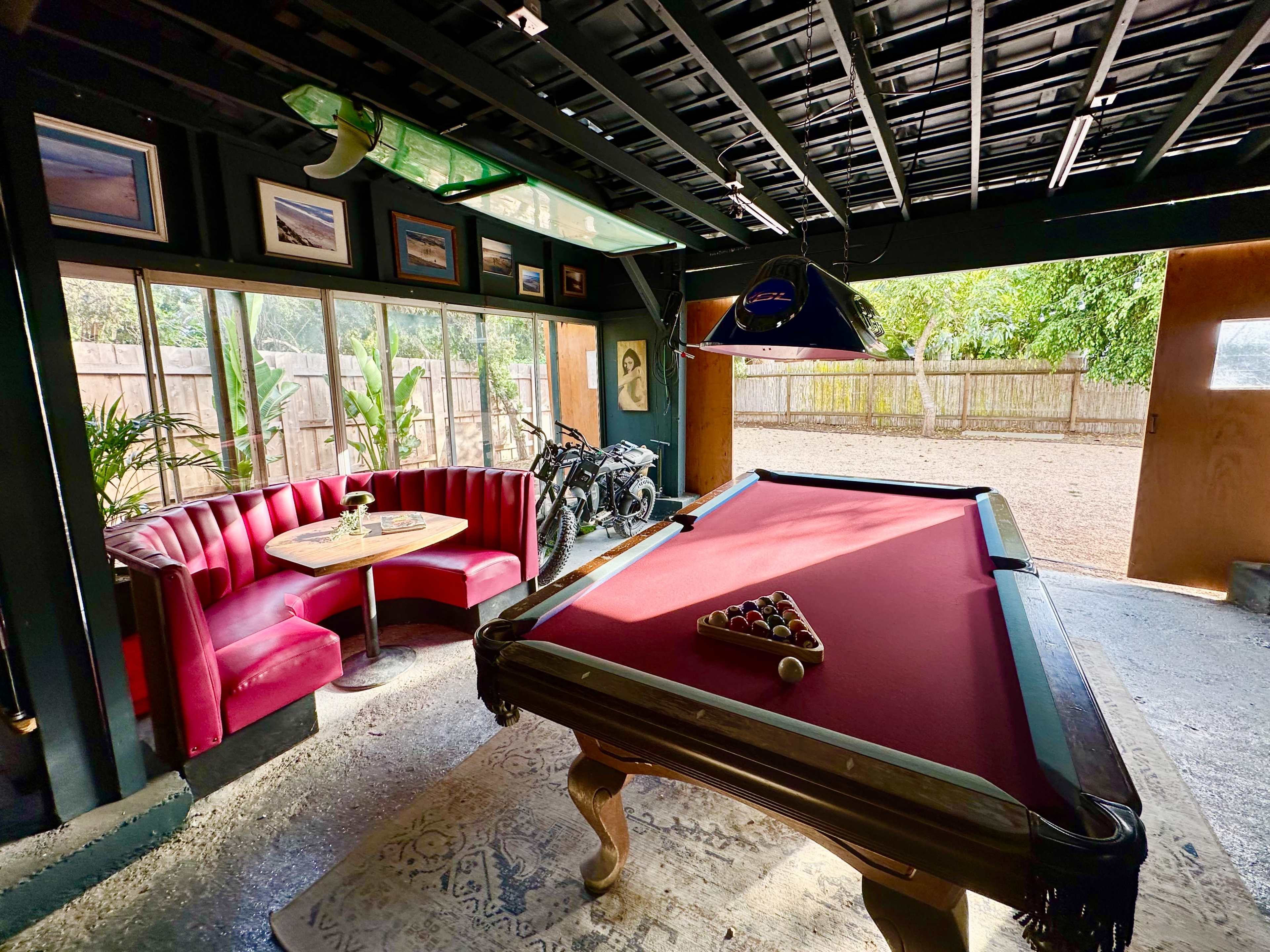 The image shows a game room featuring a red pool table, a round table with chairs, framed pictures on the walls, and a view of a garden outside through large windows.