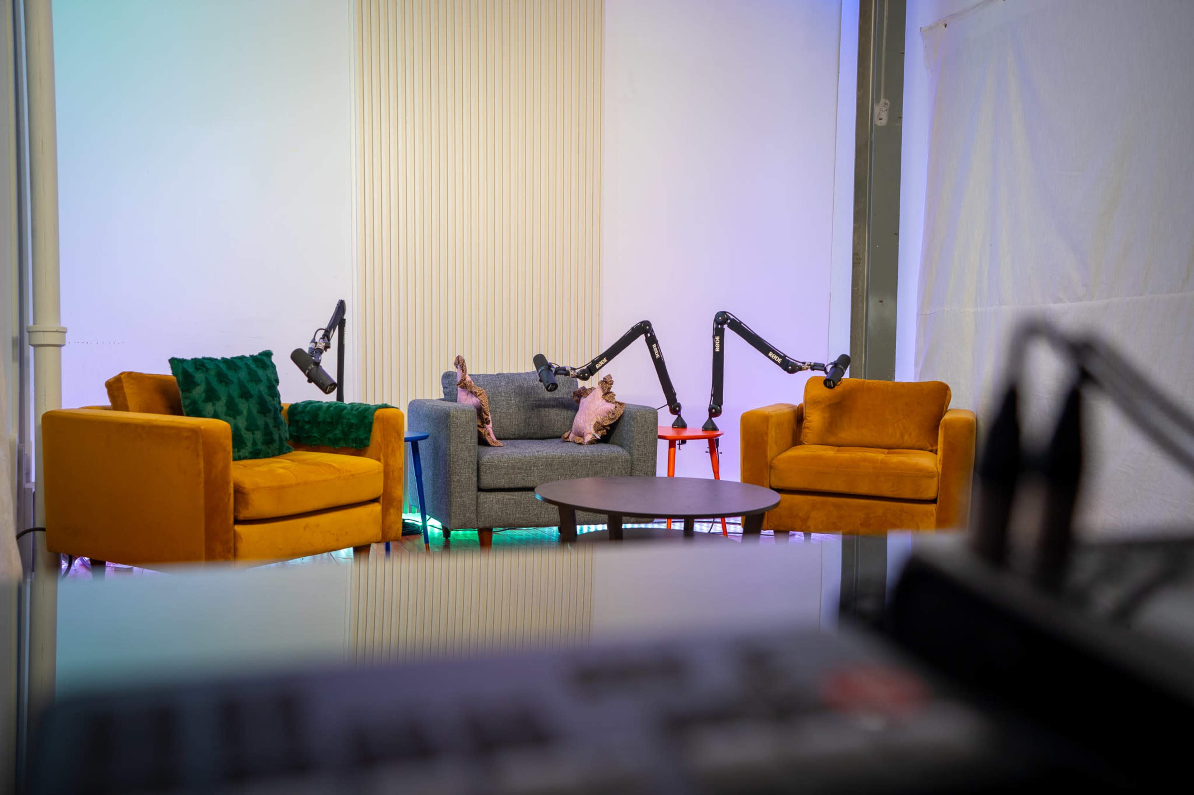 The image shows a cozy studio setup with two colorful sofas, a gray armchair, and a coffee table, illuminated by ambient lighting.