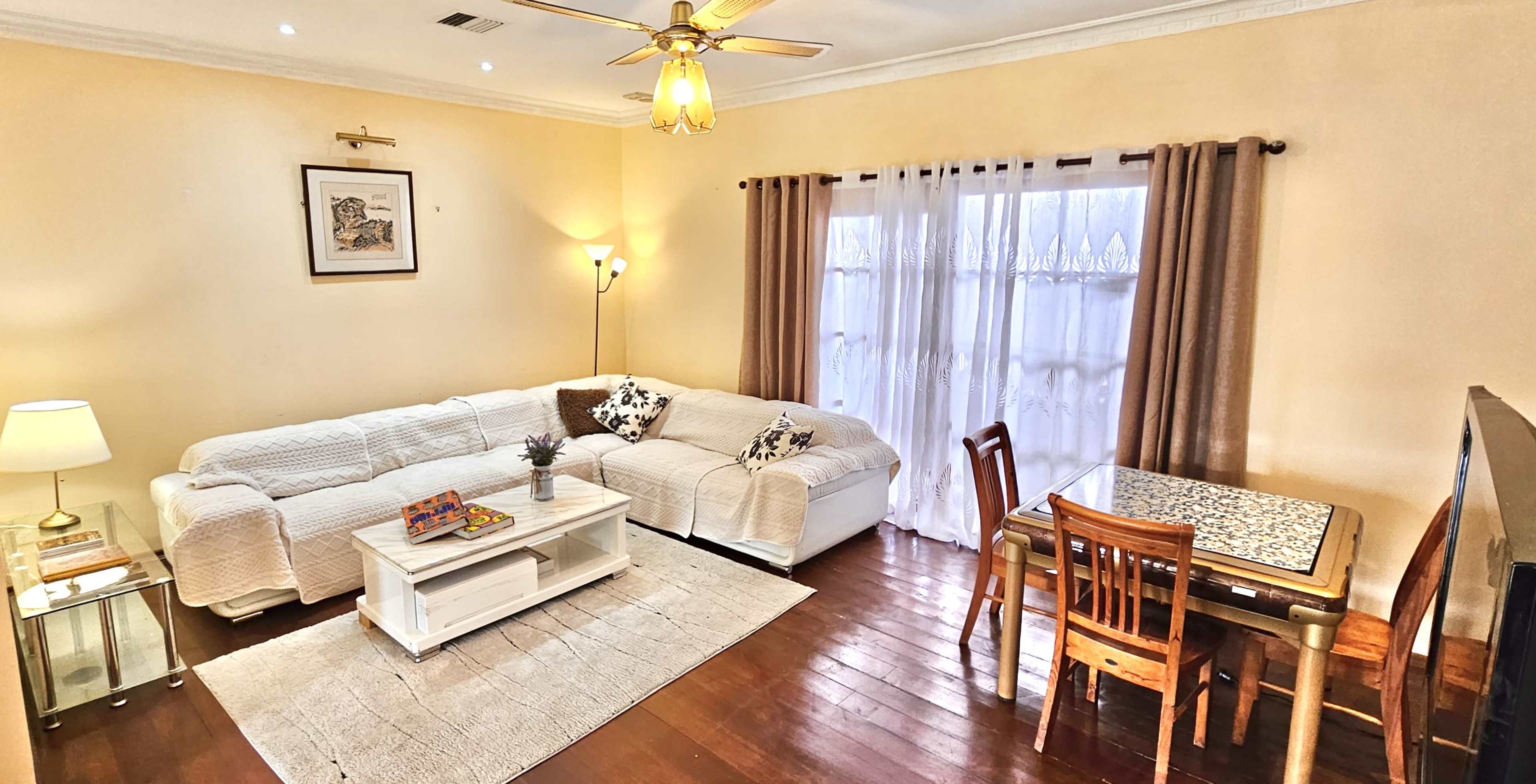 The image shows a cozy living room with a white sectional sofa, a coffee table, a dining table with chairs, and a window dressed with sheer curtains.