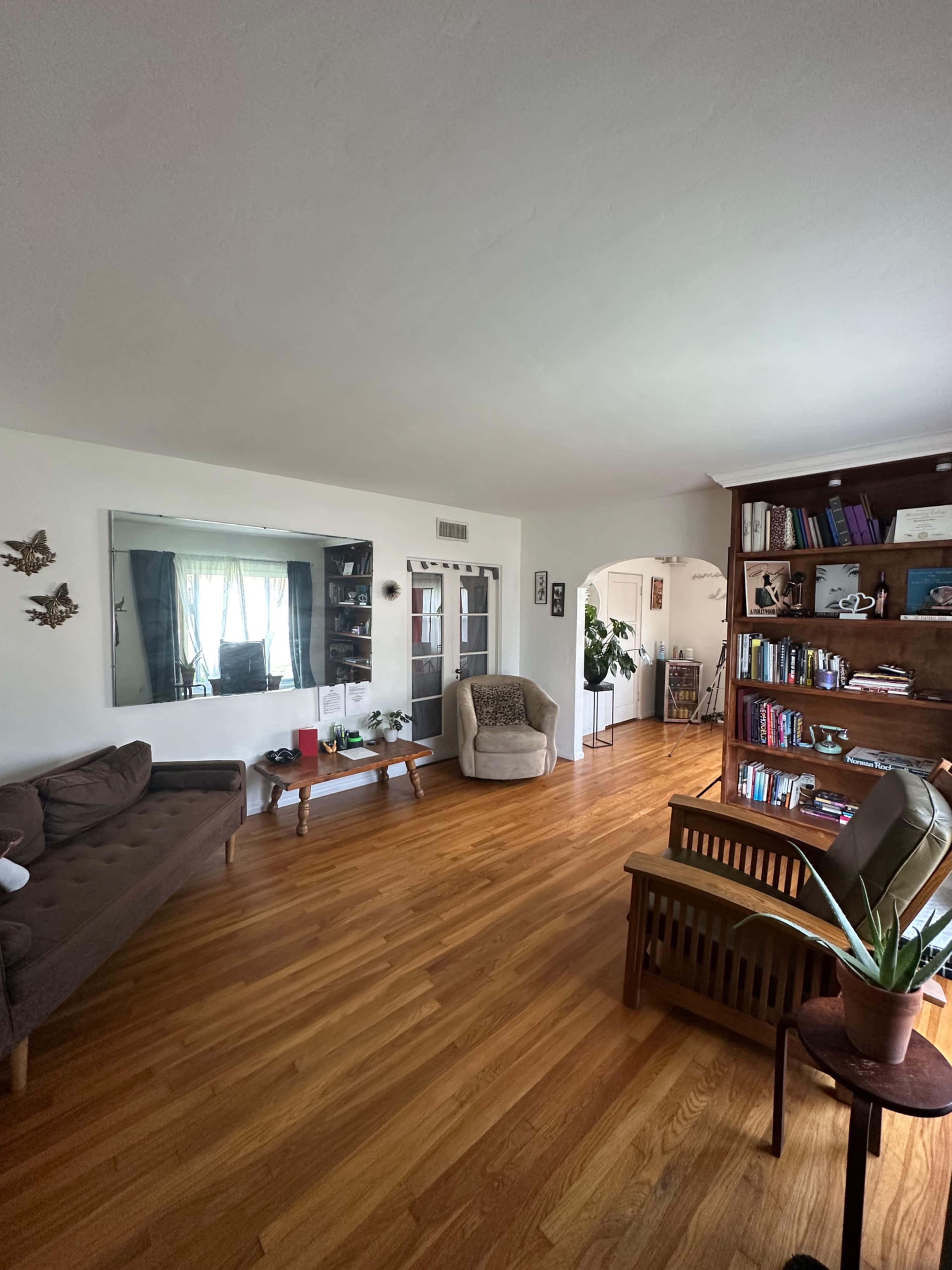 The image shows a cozy living room featuring a sofa, a bookcase filled with books, and natural light streaming through large windows.