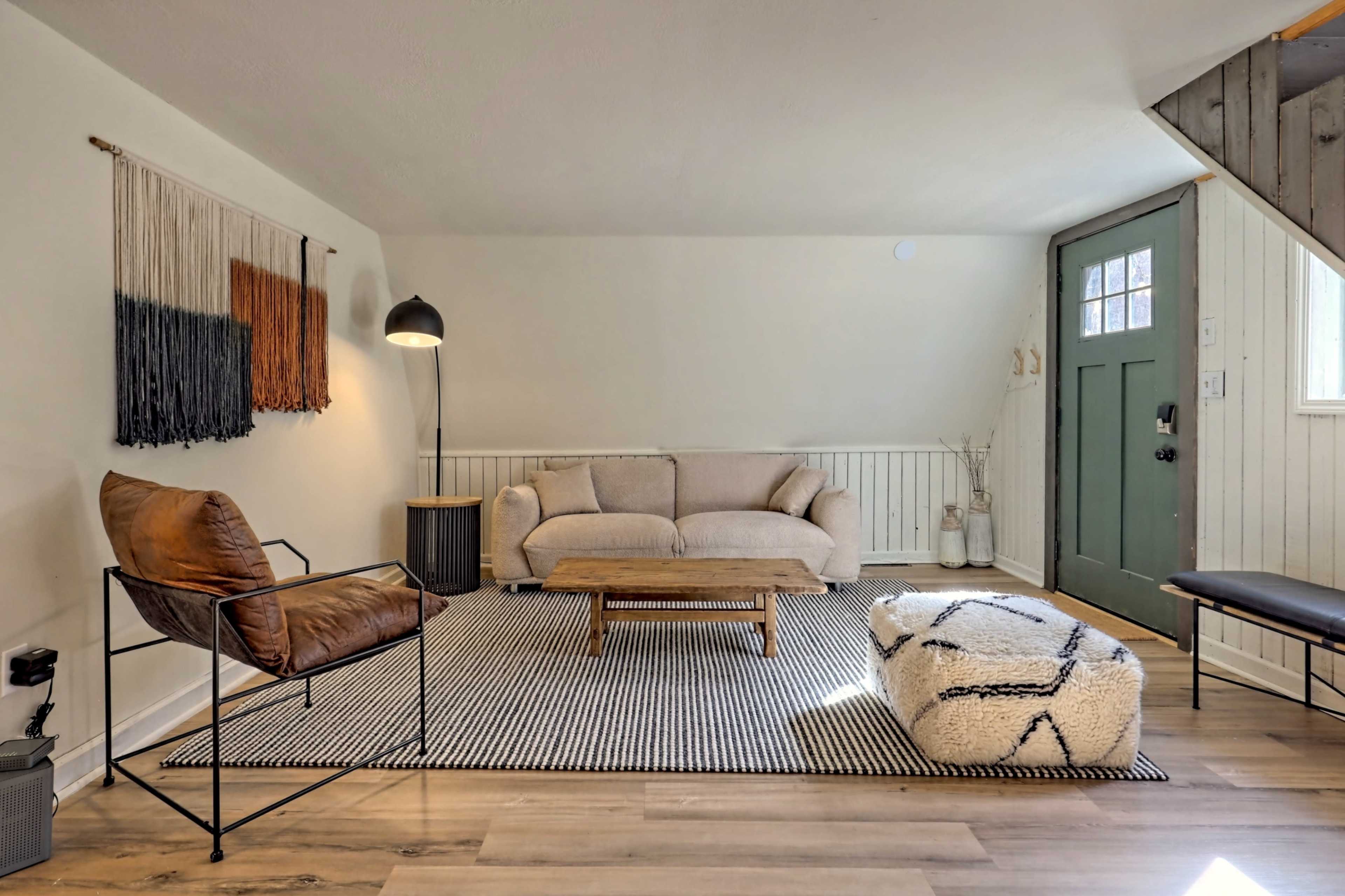 A light-filled living room features a beige sofa, a woven wall hanging, a wooden coffee table, a black and white patterned rug, and a green front door.