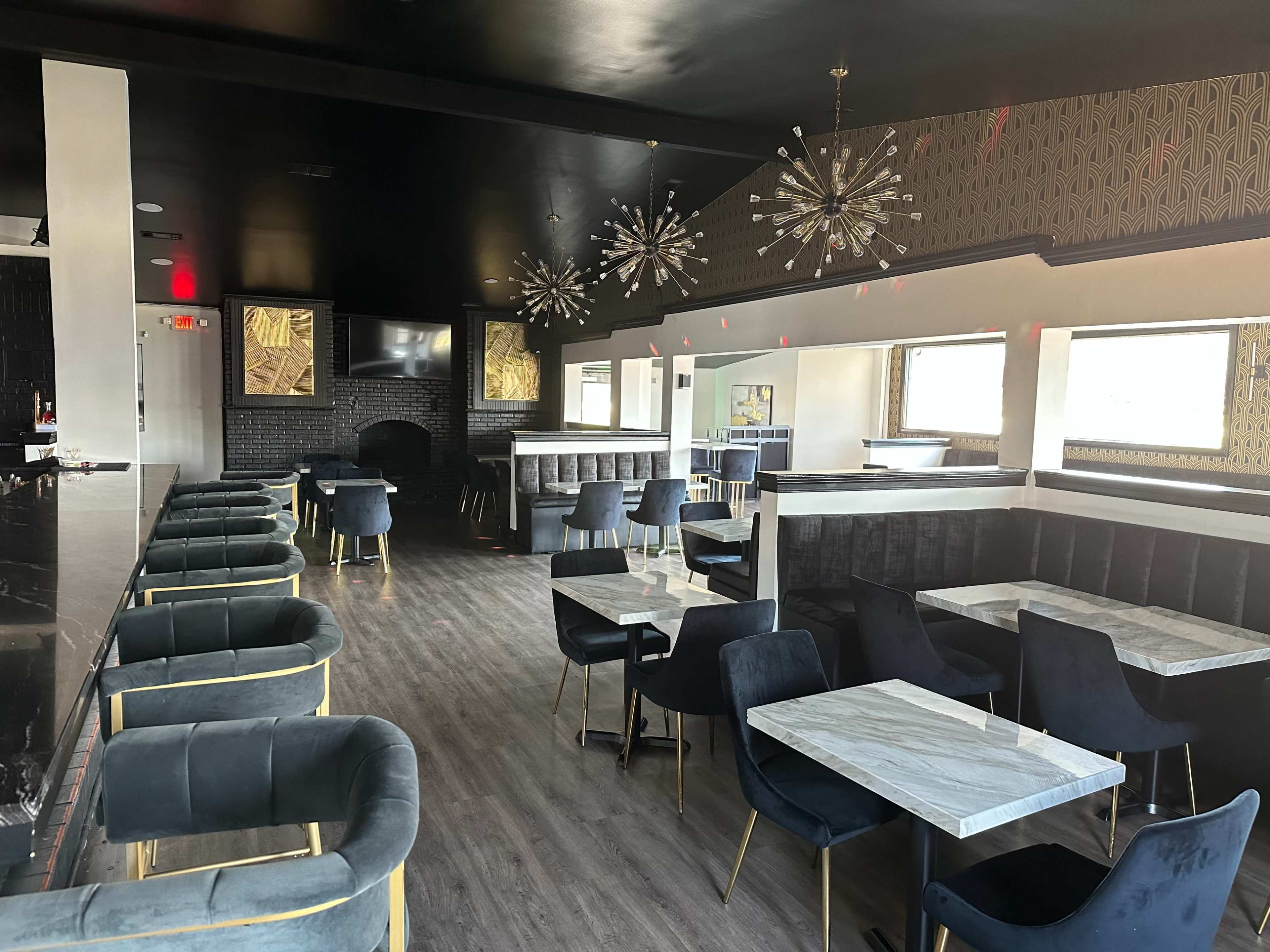 The image shows a modern restaurant interior featuring black and gold decor, dark velvet chairs, marble-topped tables, and hanging light fixtures.