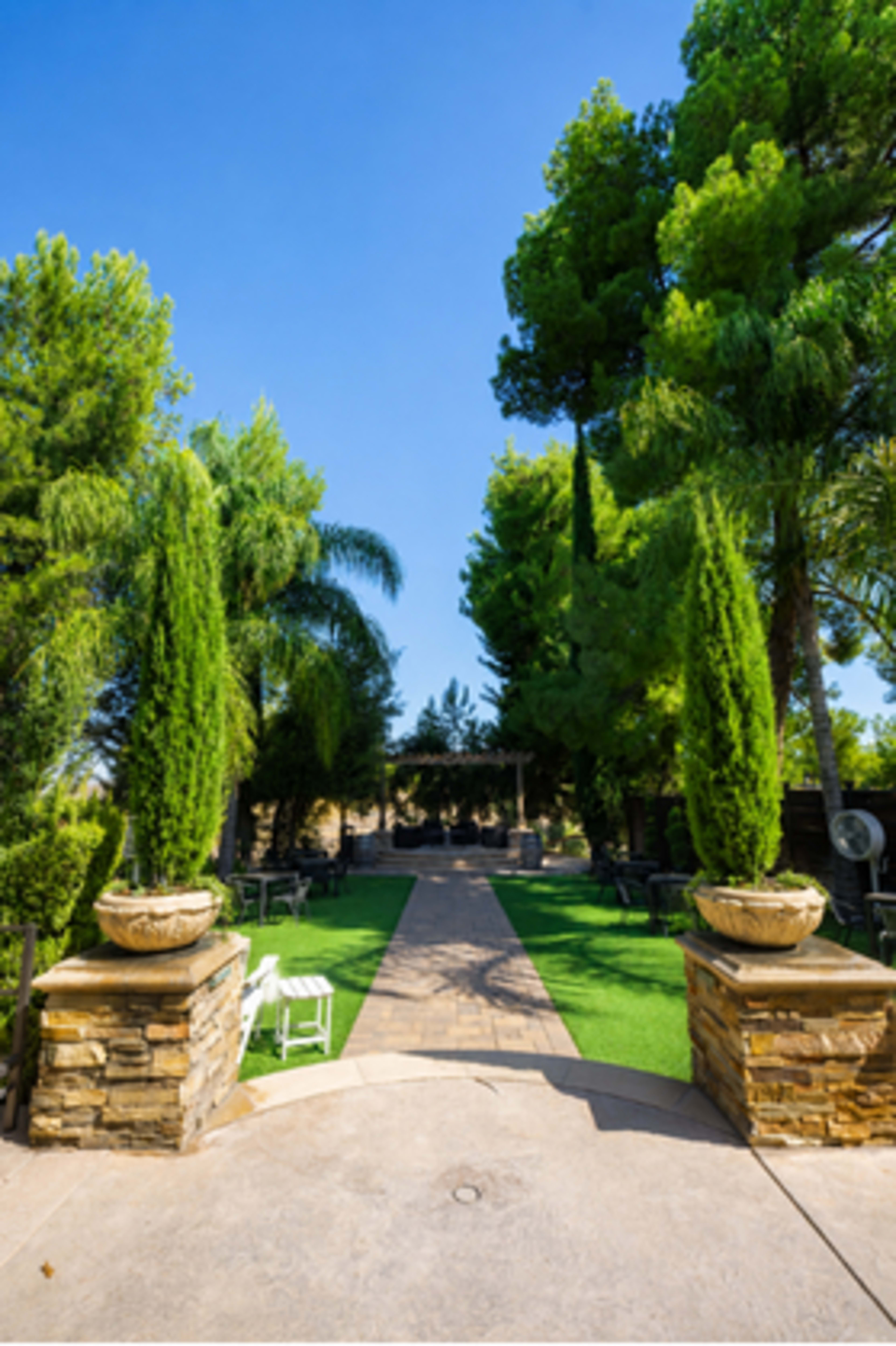 A stone pathway leads through a landscaped garden with tall trees and decorative planters on either side.