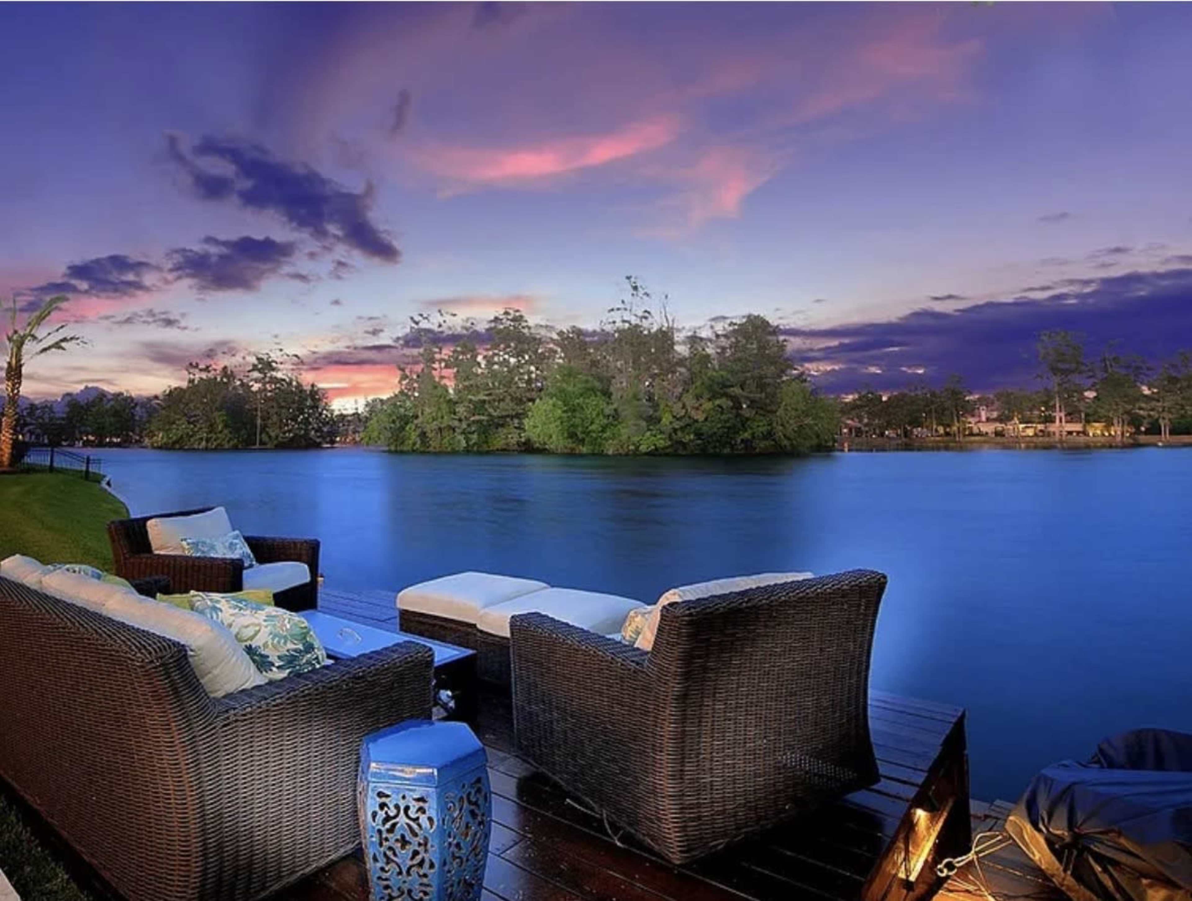 A seating area with wicker furniture is positioned beside a calm body of water, under a colorful sky at dusk.