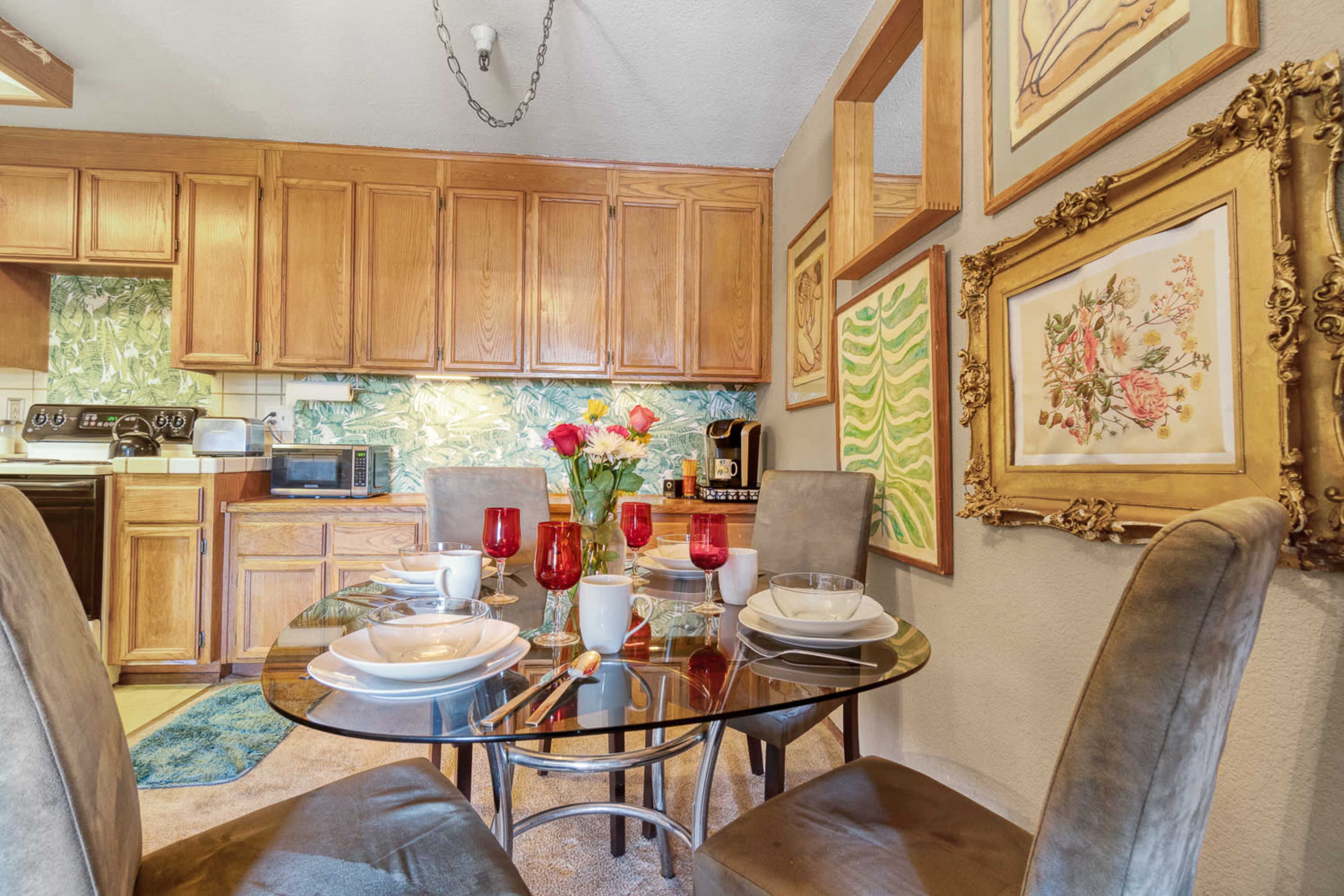 A dining area features a glass table set with dishes and flowers, adjacent to a kitchen with wooden cabinets and decorative wall art.