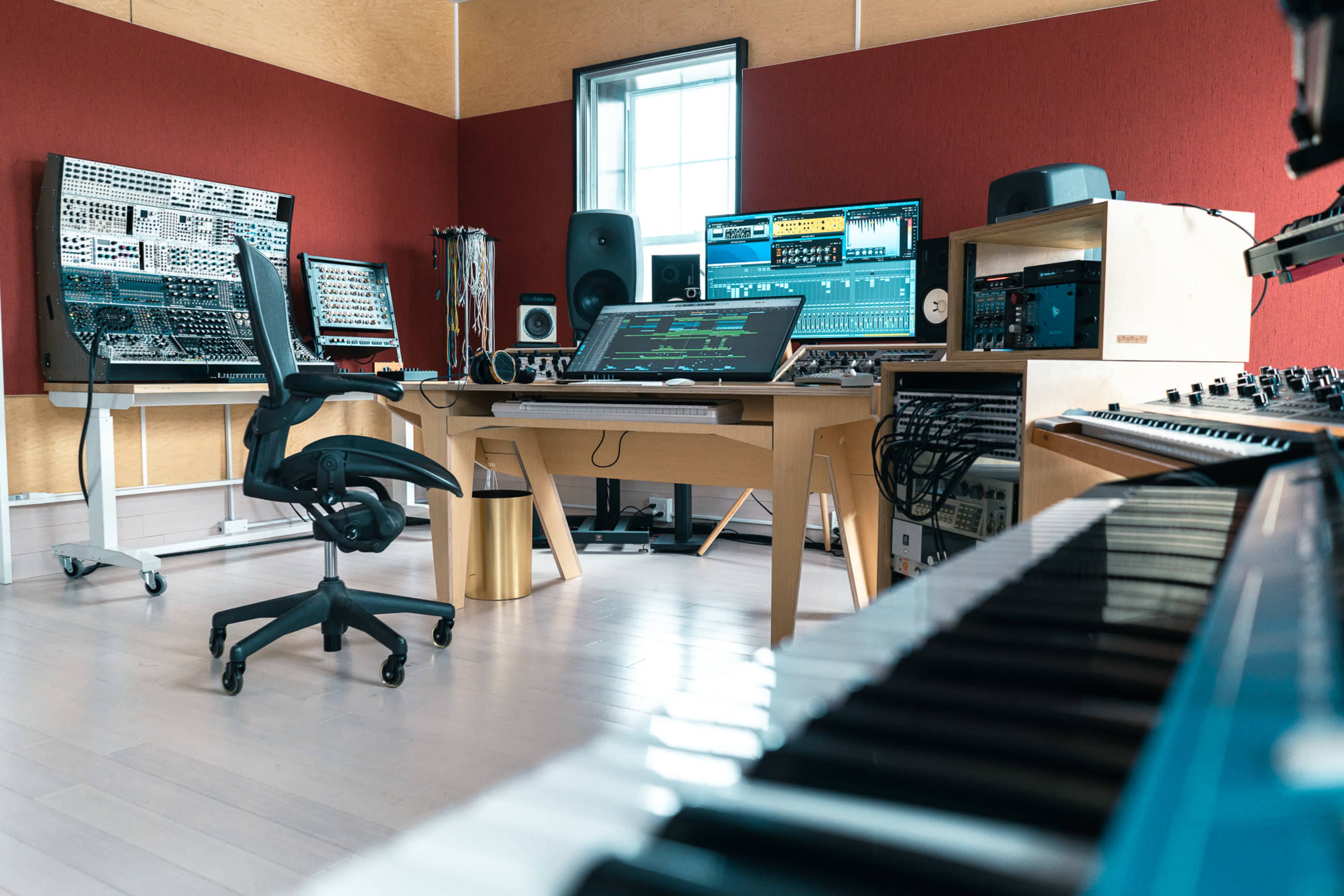 A modern music studio features multiple synthesizers, a large computer workstation, an arranged keyboard, and sound equipment amidst a minimalist interior.