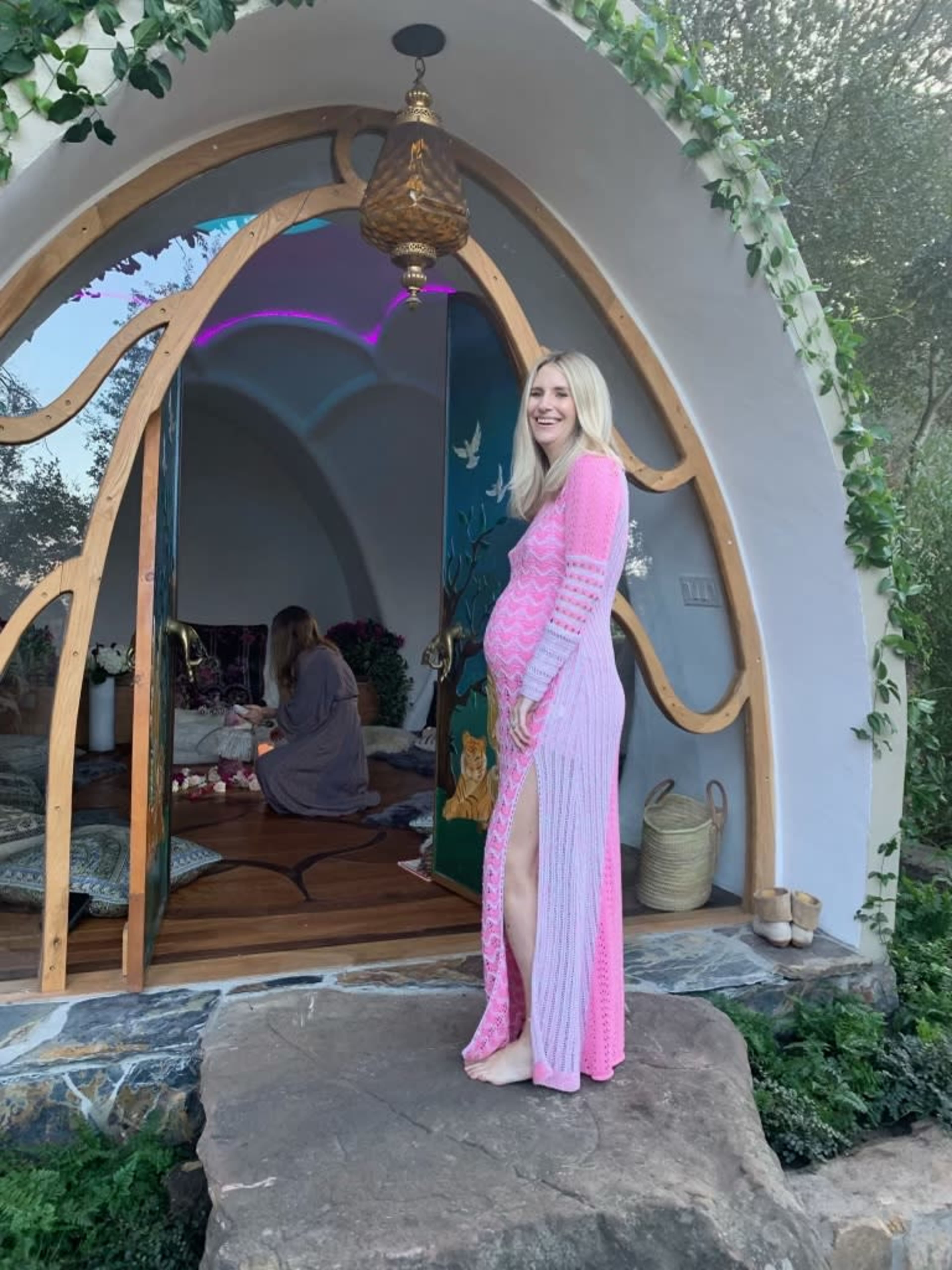 A woman in a pink and white striped dress stands beside an arched entrance with greenery, while another person is seated inside the cozy, decorated space.