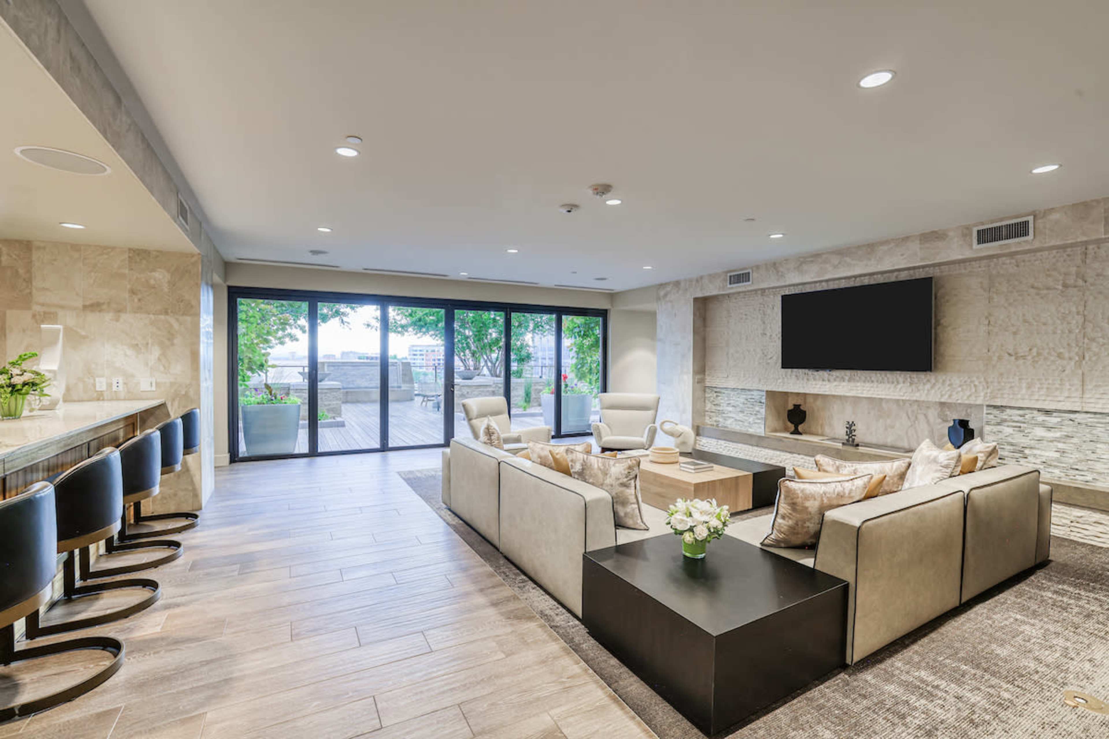 The image shows a modern living area featuring a sectional sofa, a coffee table, and large windows that open to a deck with greenery.