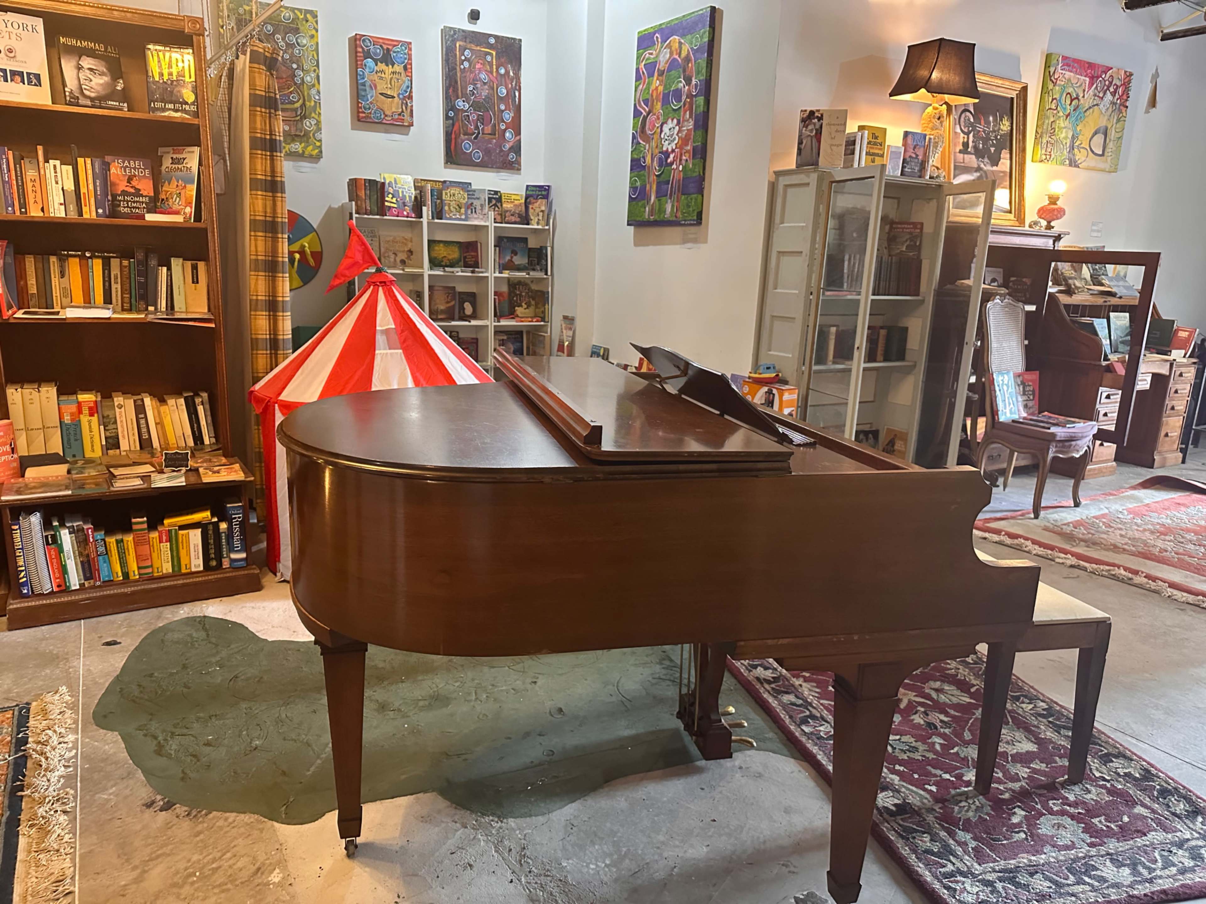 A wooden grand piano is positioned in a cozy room filled with bookshelves, colorful artwork, and a decorative circus-themed tent.