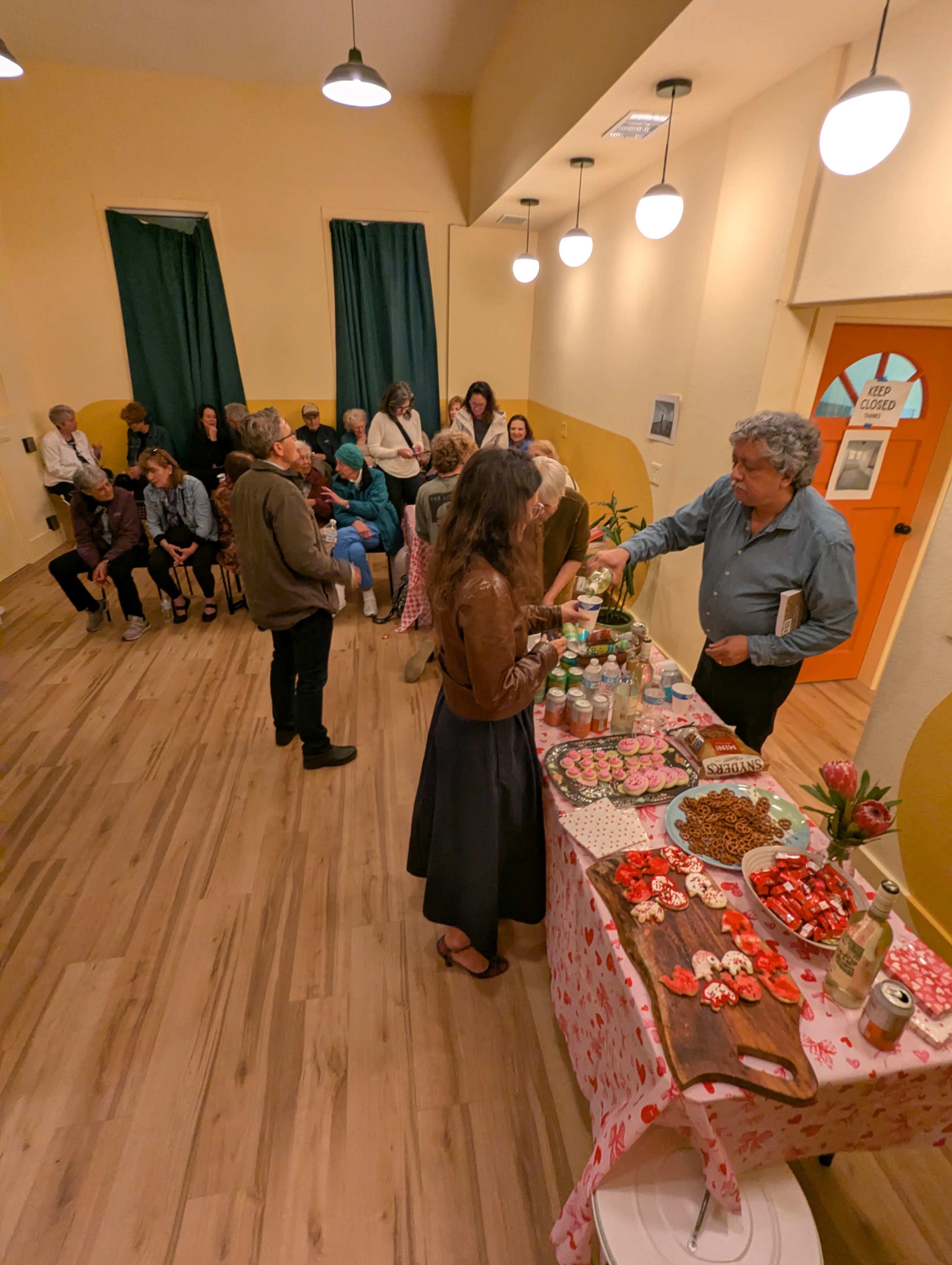 A diverse group of people gathers in a warmly lit room, some seated while others converse around a table filled with various treats and beverages.