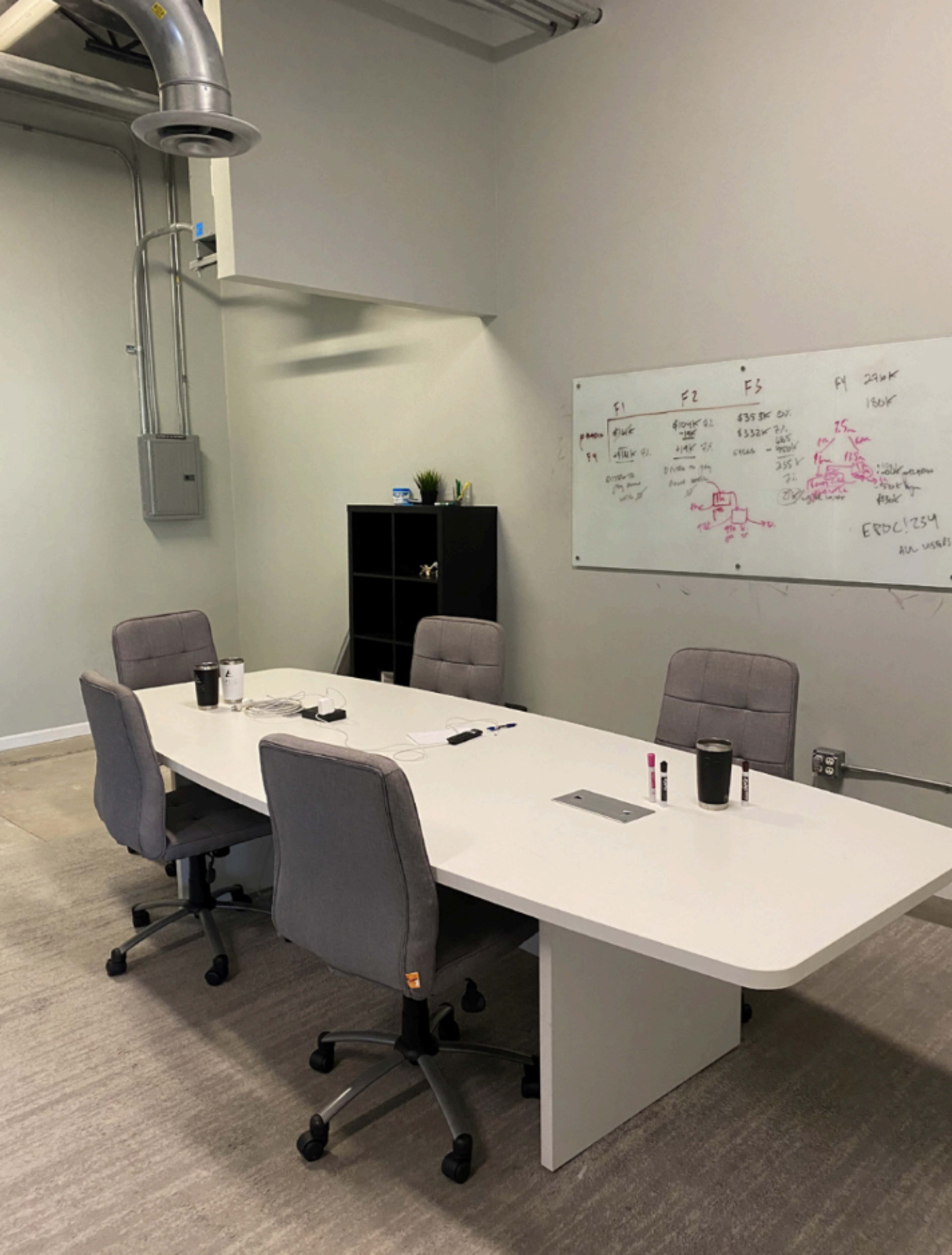 A modern conference room features a long white table surrounded by four gray chairs, with a whiteboard displaying notes and a plant on a nearby shelf.