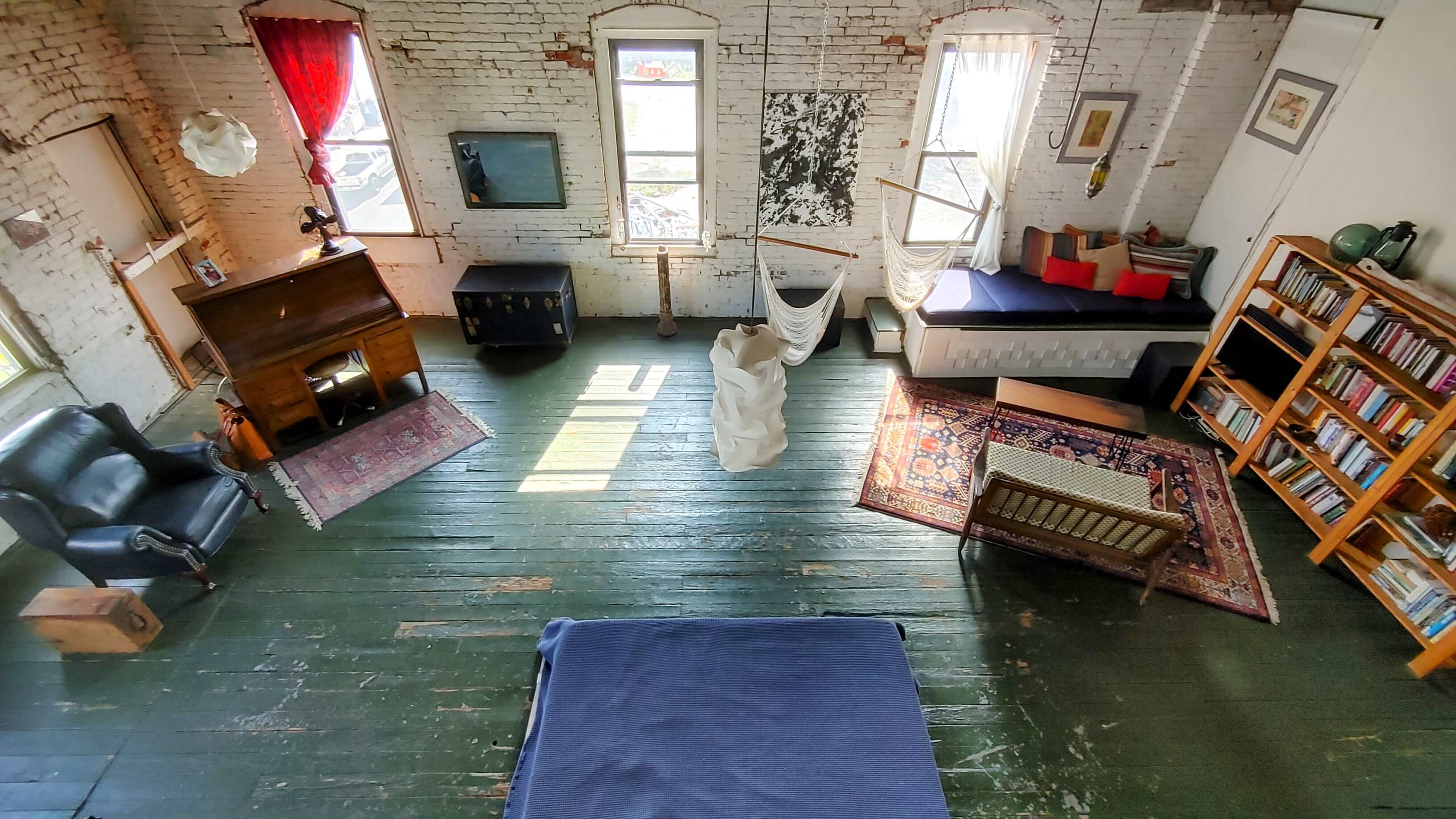 The image shows a spacious, rustic room with wooden floors, large windows letting in light, and various furniture pieces including a bed, a chair, and a bookshelf.