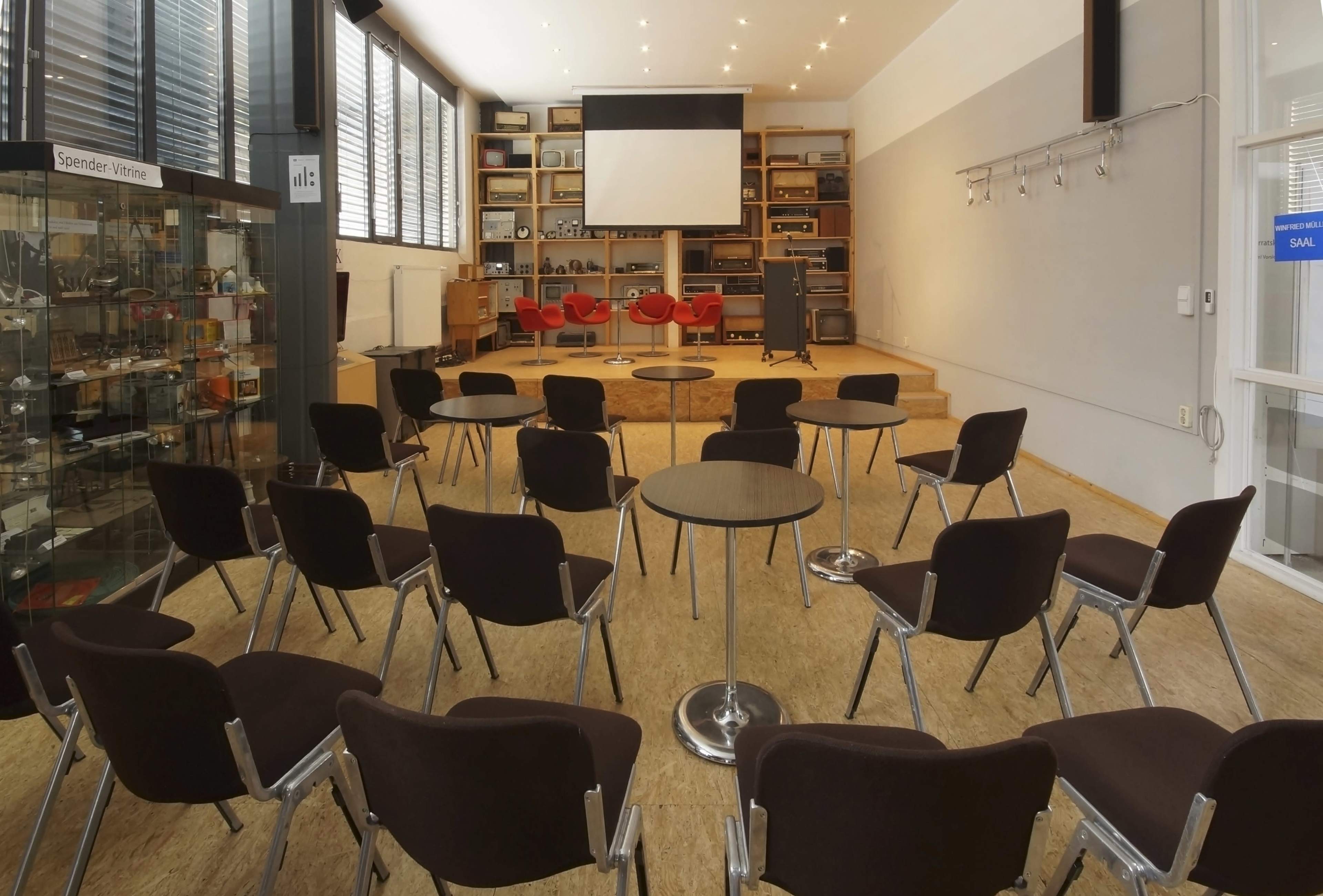 A meeting room with rows of chairs facing a stage and a large screen at the front.