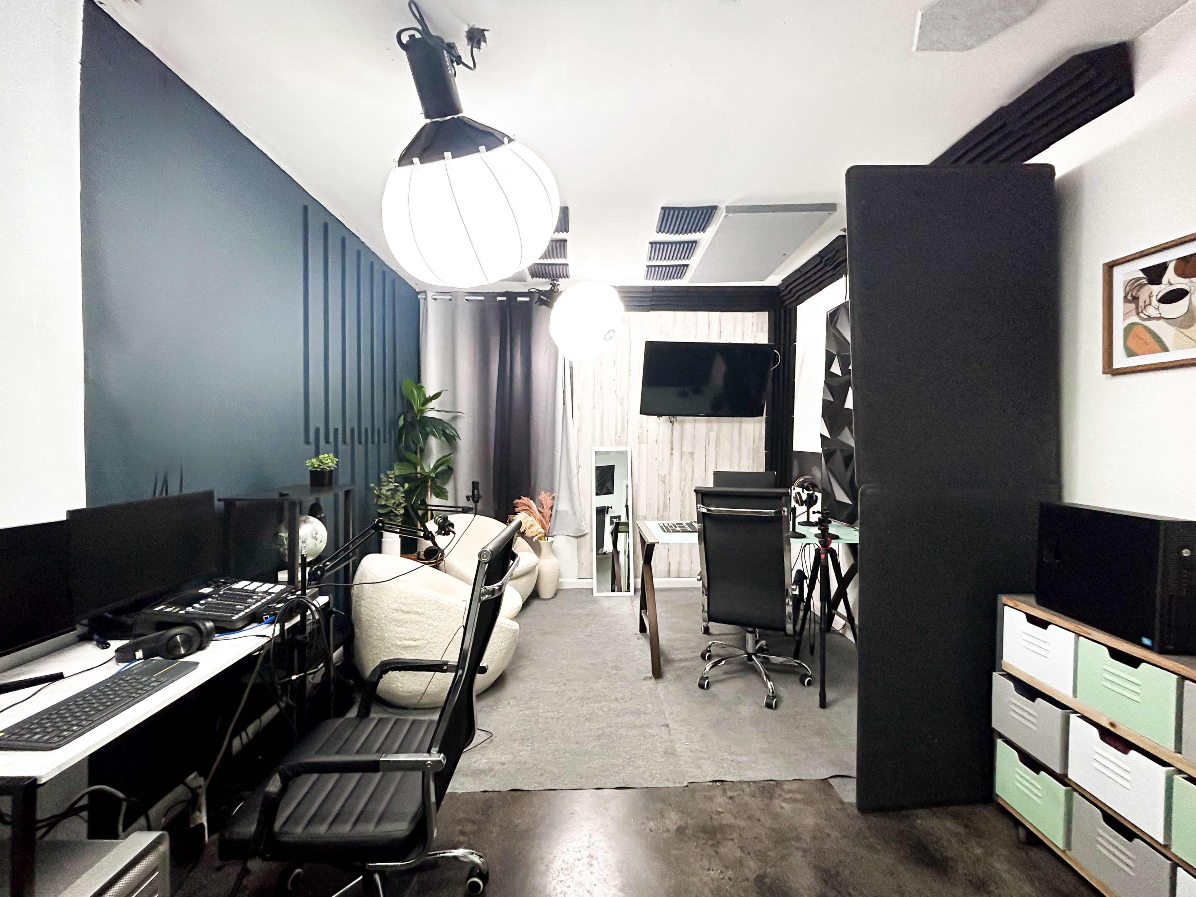 A modern office space with multiple workstations, a large light fixture, and some plants, separated by a partition.