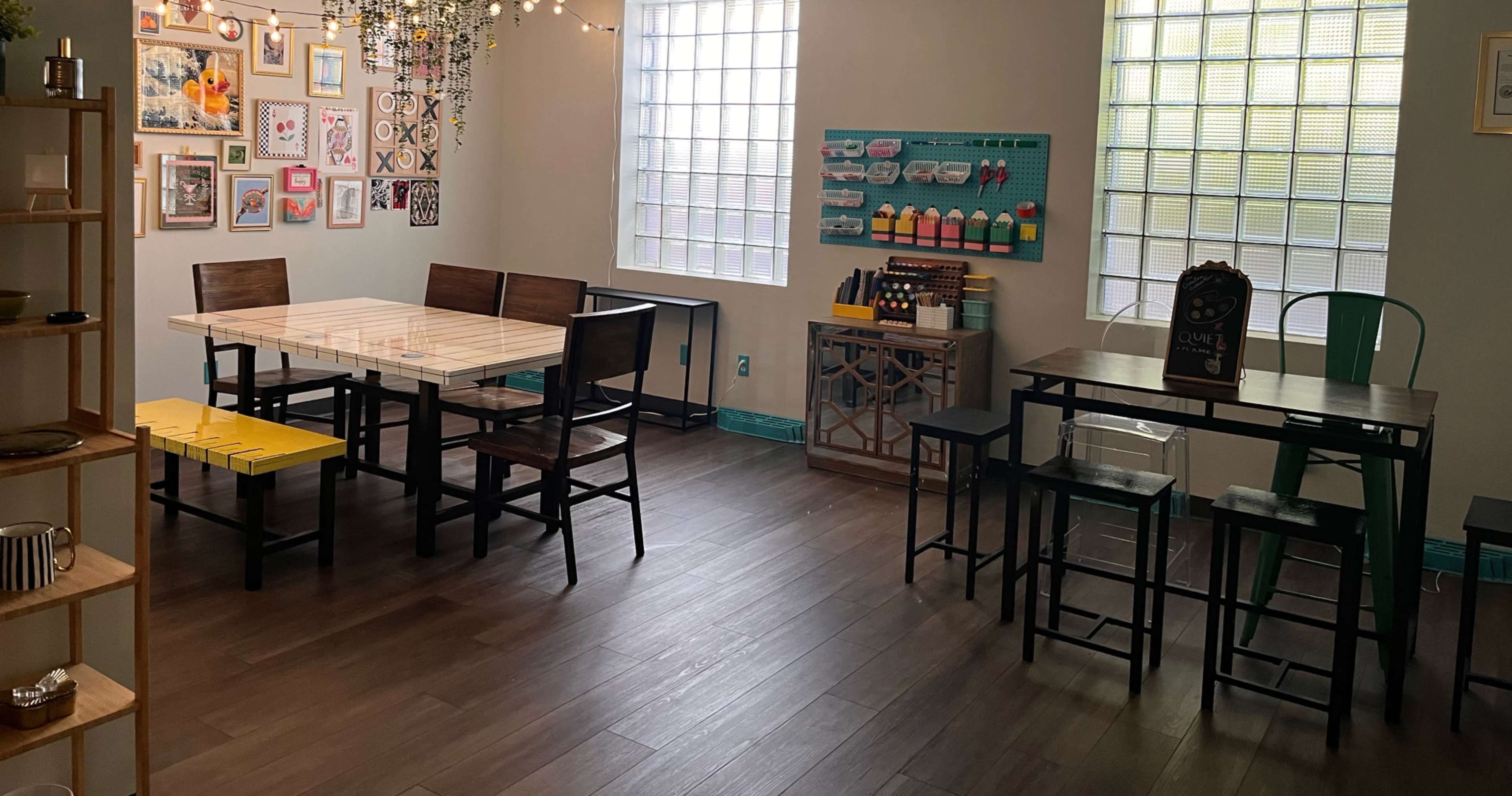 The image shows a brightly lit interior space with two tables, a gallery wall of framed pictures, and a variety of seating arrangements.