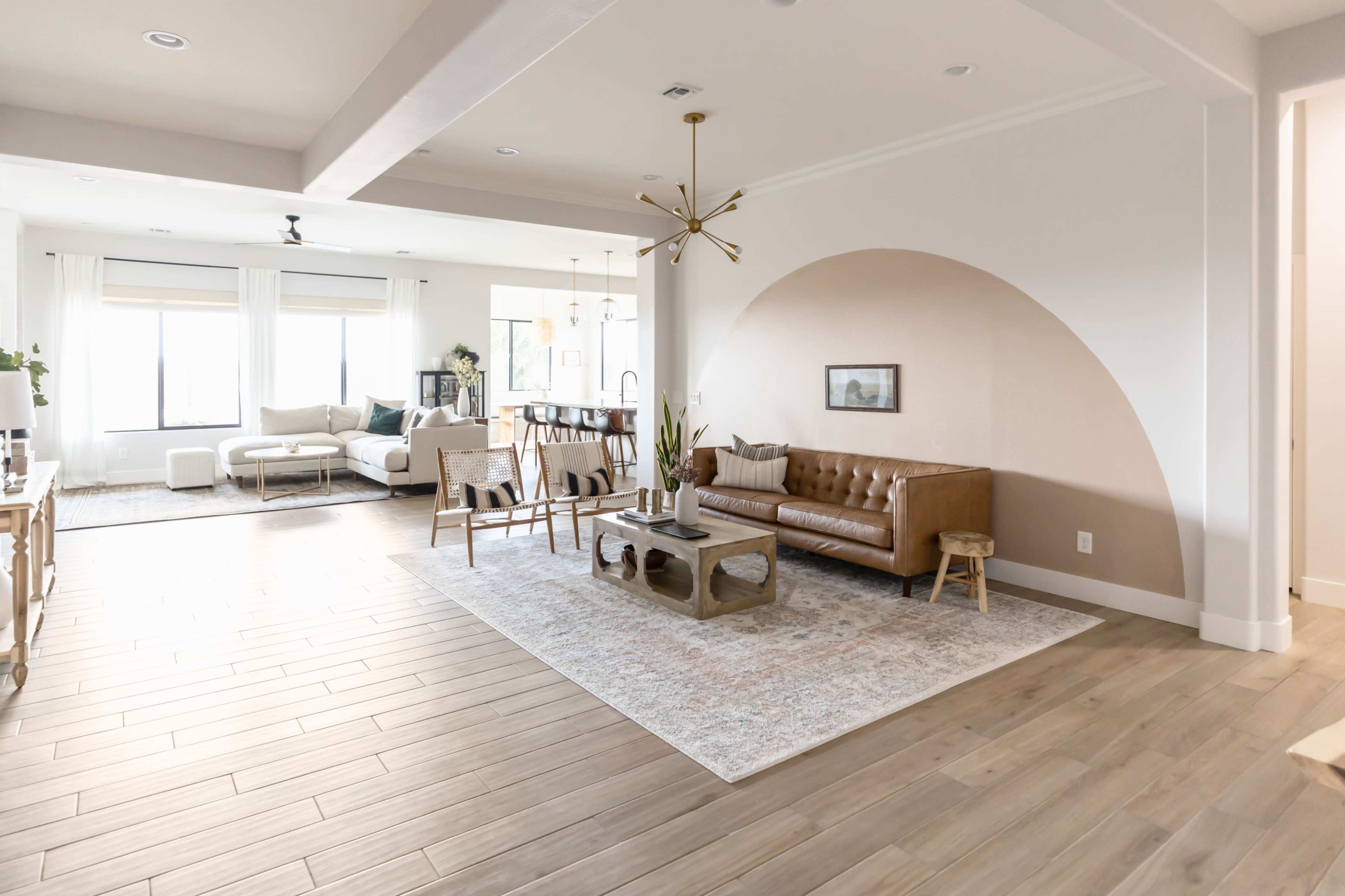 A modern living room with a brown leather sofa, a coffee table, and an area rug, featuring large windows and an open layout connecting to an adjacent space.