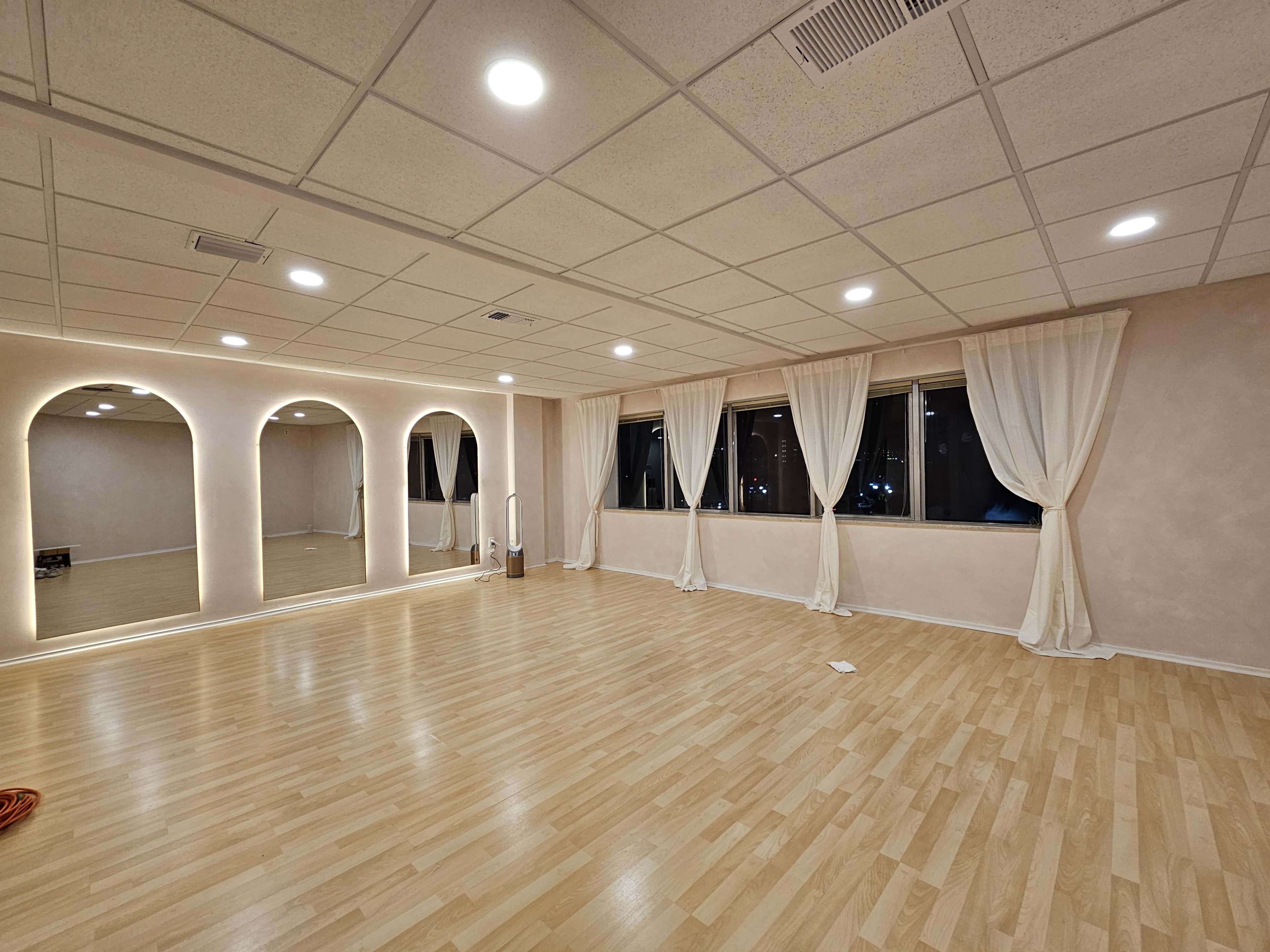 The image shows an empty room with wooden flooring, large windows, and mirrored arches on one wall, illuminated by ceiling lights.