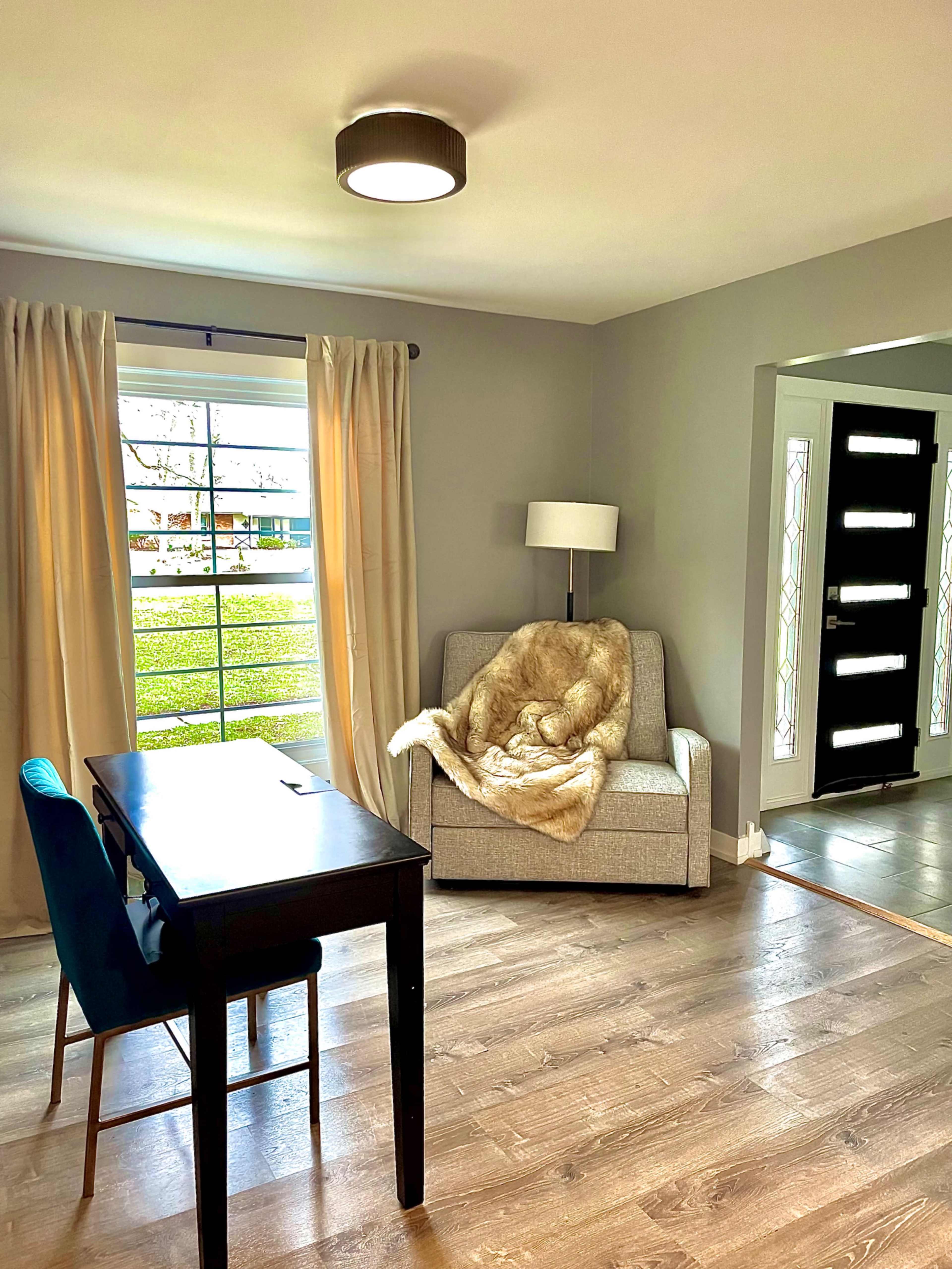 The scene shows a living room with a gray armchair draped in a light-colored blanket, a wooden desk with a blue chair, and a modern front door featuring large glass panels.