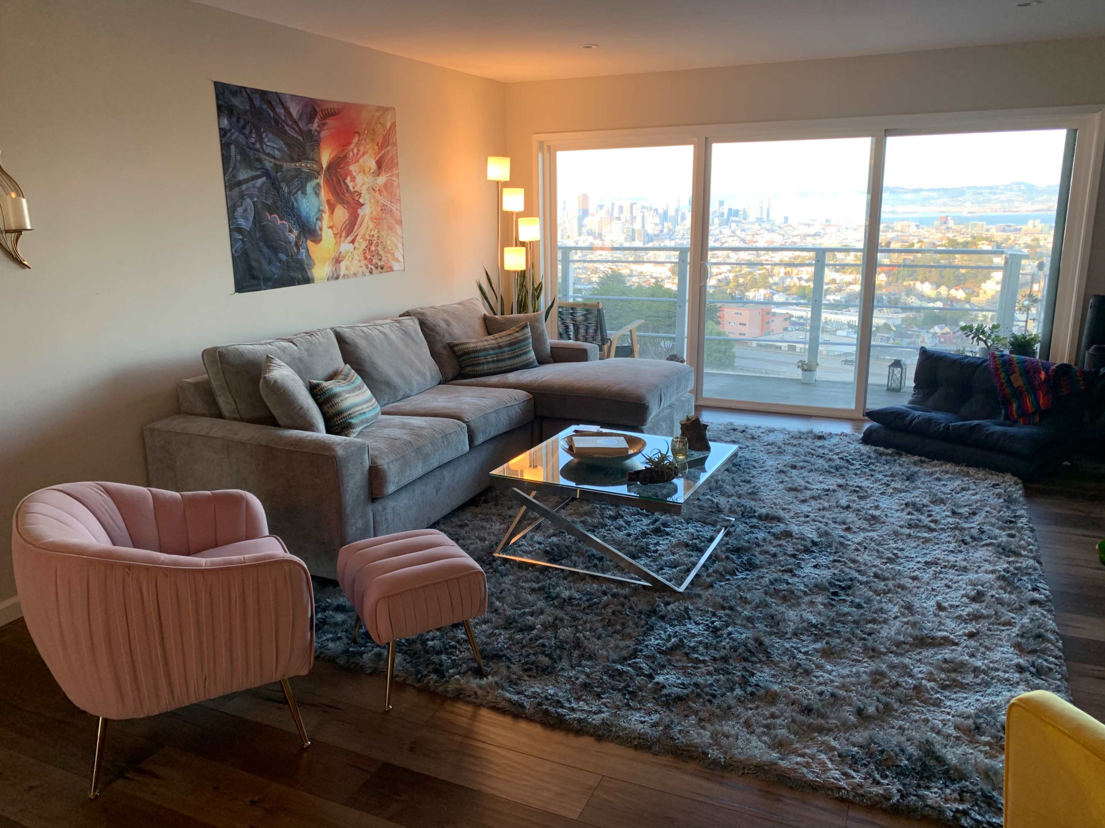 A modern living room features a gray sectional sofa, a pink armchair, a glass coffee table, and large windows showcasing a city skyline.