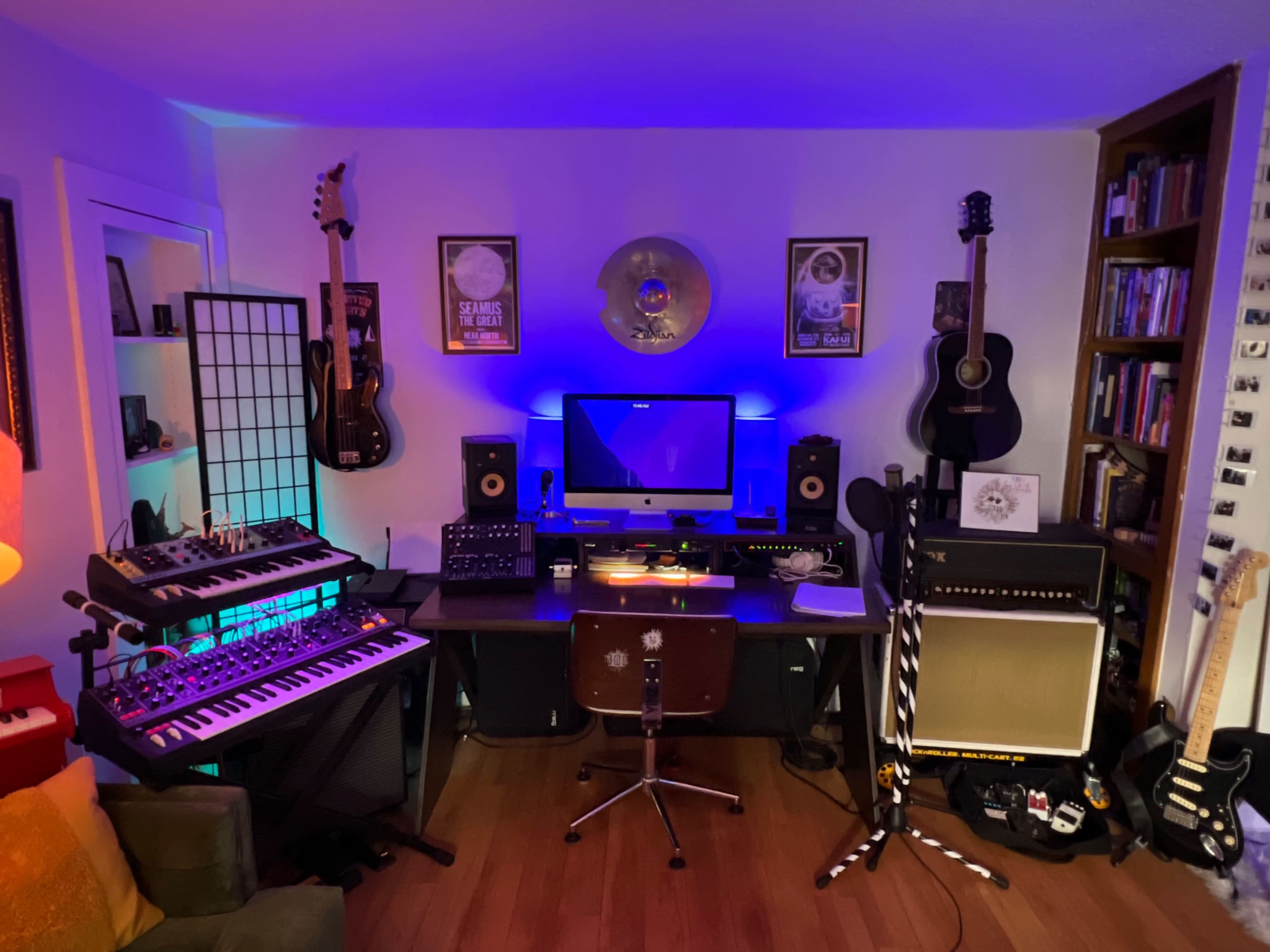 The image shows a music studio setup with a computer workstation, synthesizers, guitars, and various audio equipment arranged in a brightly lit room.