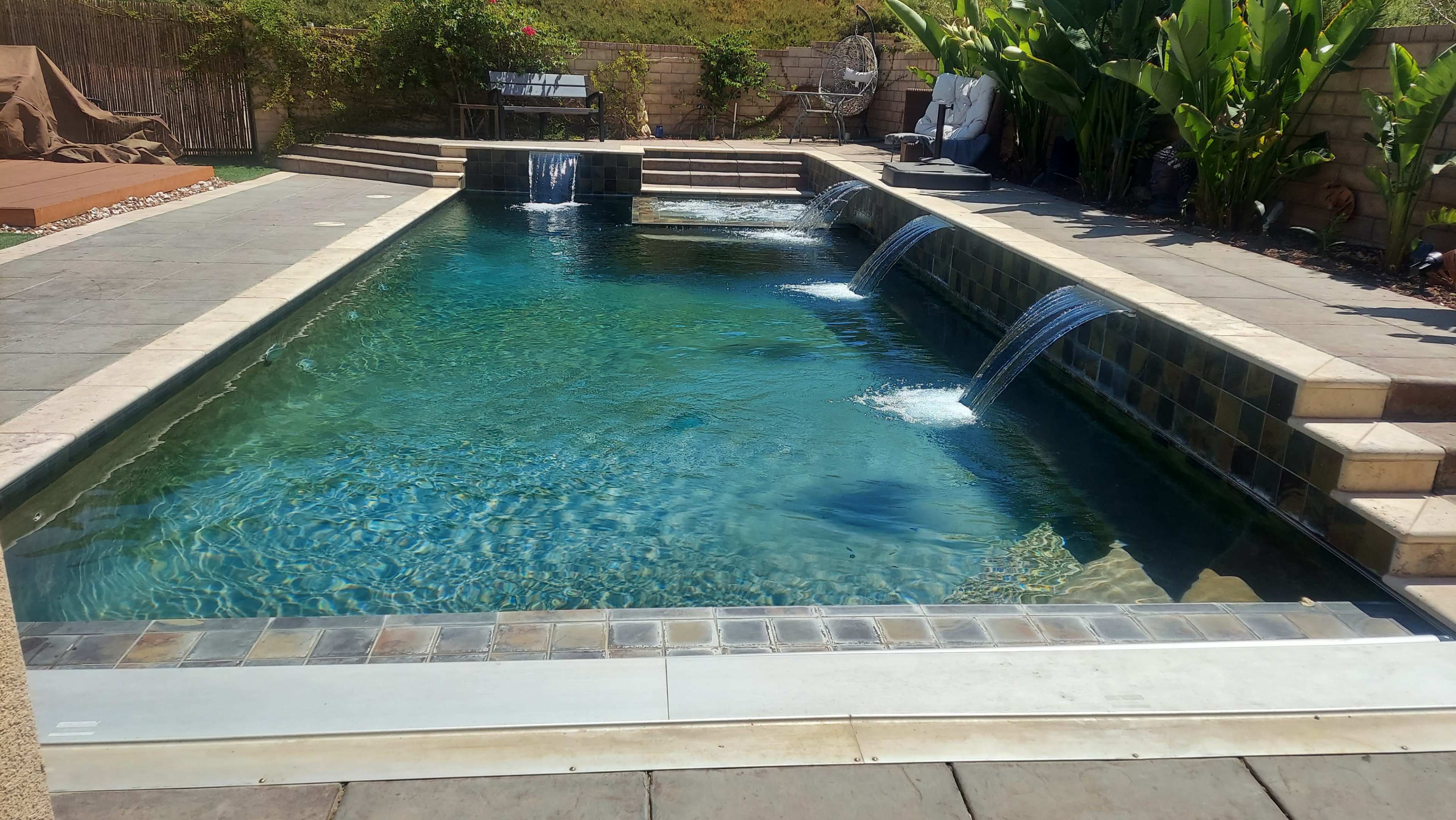 The image features a rectangular swimming pool with three water features creating waterfalls, surrounded by stone decking and lush greenery.