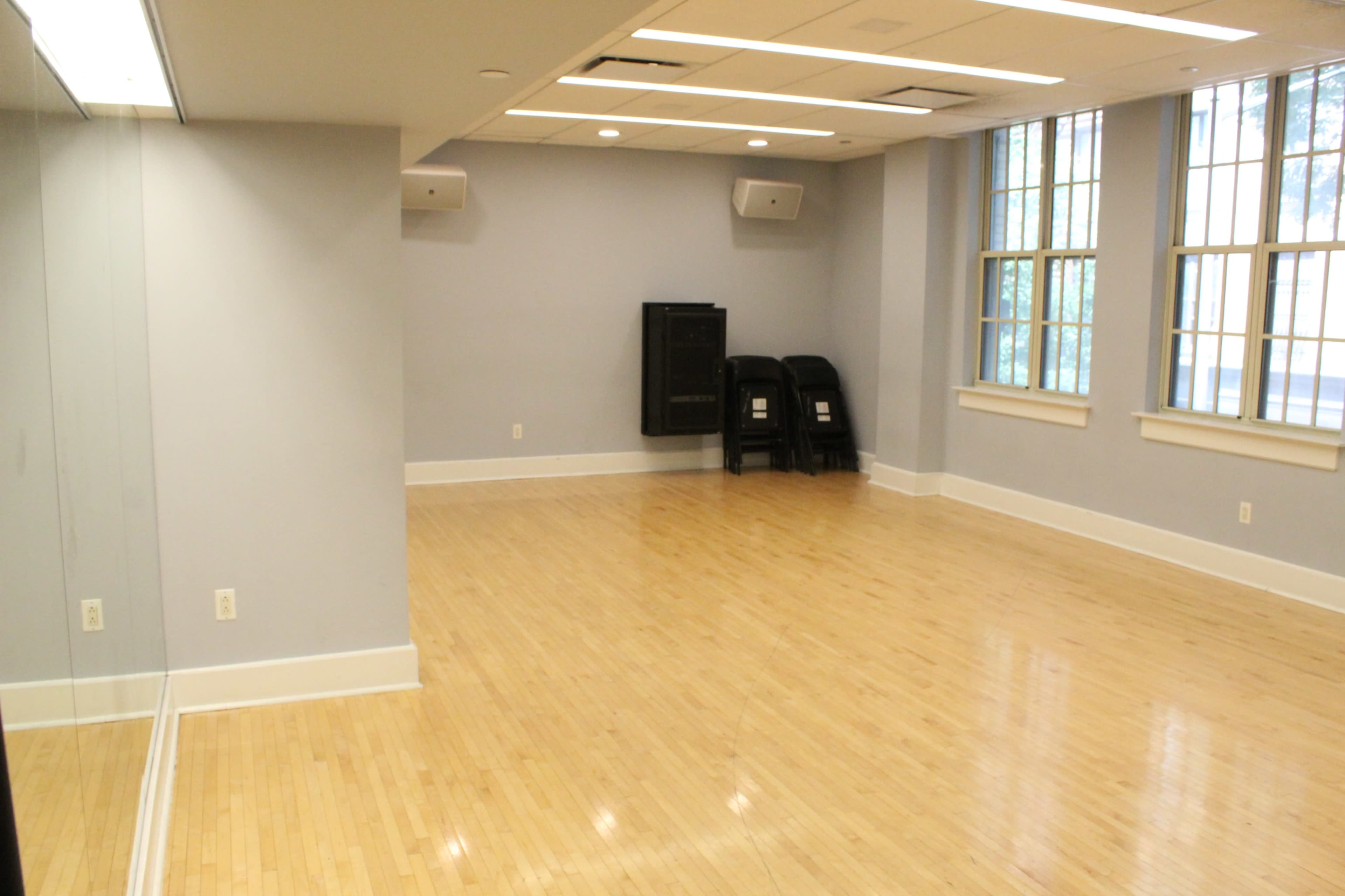 The image shows a spacious, empty room with wooden floors, large windows, and a mirrored wall on one side.