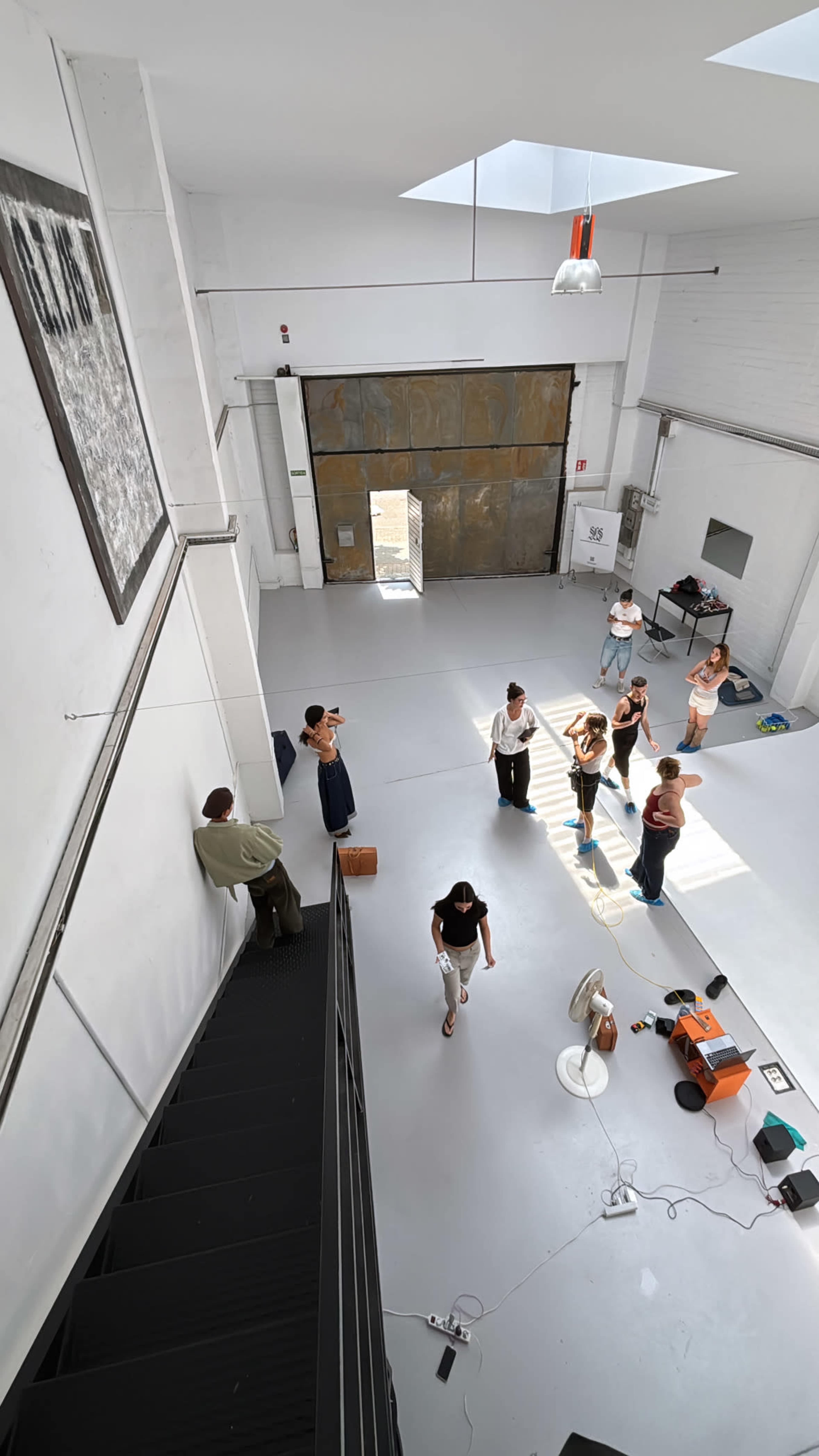 A group of people is gathered in a spacious, well-lit room with a large open door, while some are standing near a staircase and others are engaged in conversation or activity on the floor.