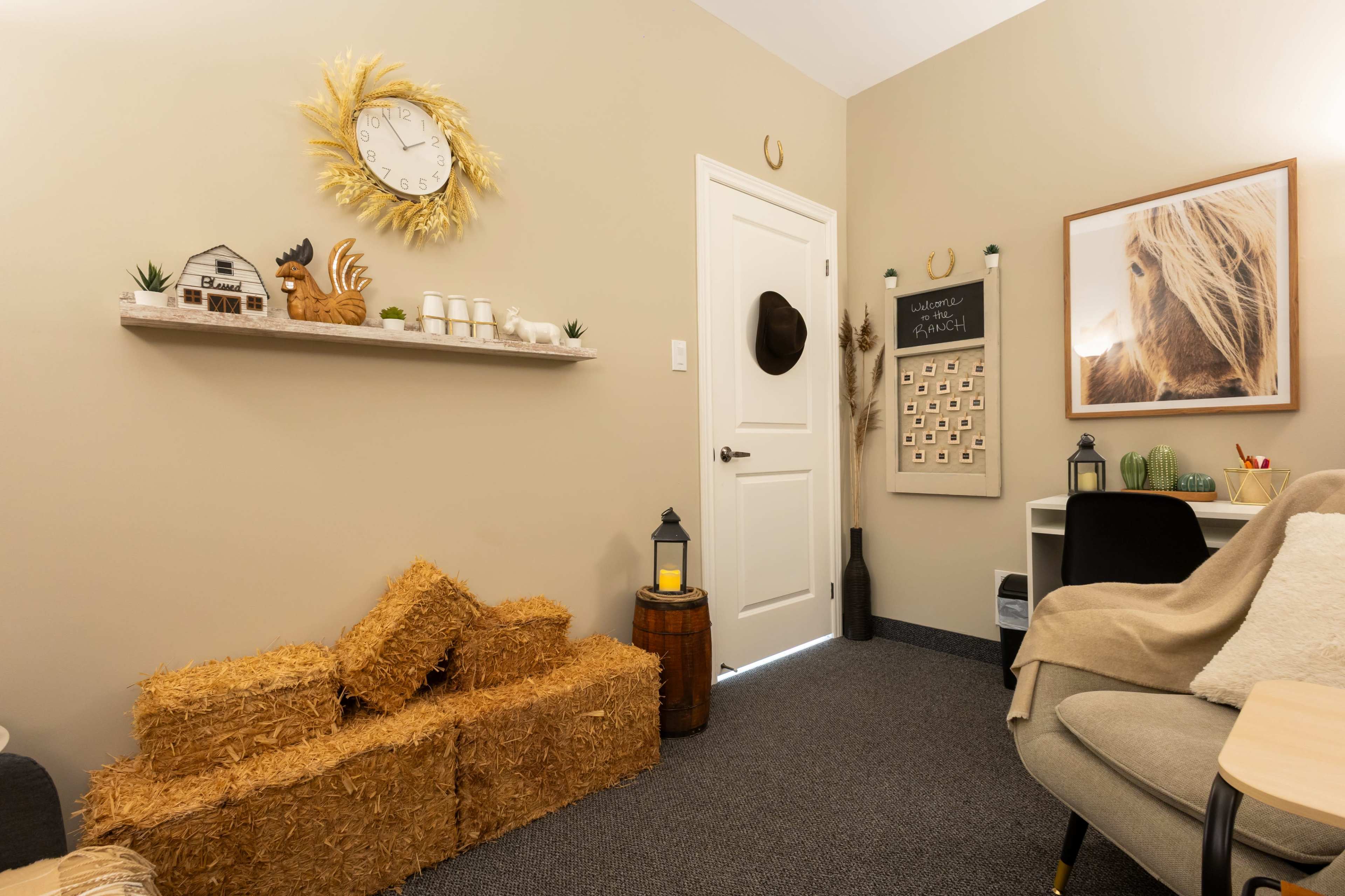 The image shows a cozy room featuring straw bales, a clock, a small decorative shelf with a house and rooster figurine, and a desk with a chair.