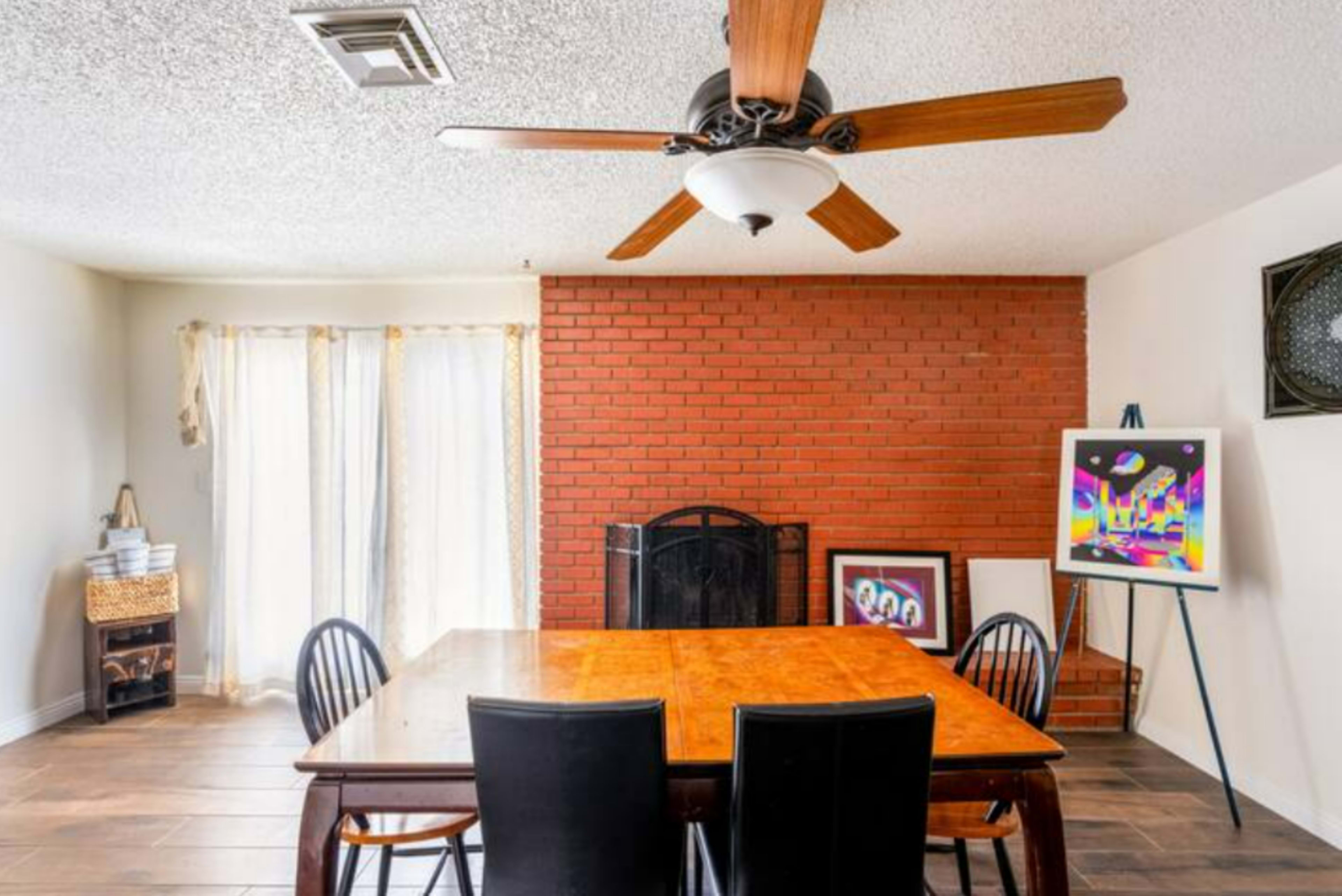 A dining area with a wooden table surrounded by black chairs, a brick wall with a fireplace, and large windows with sheer curtains.