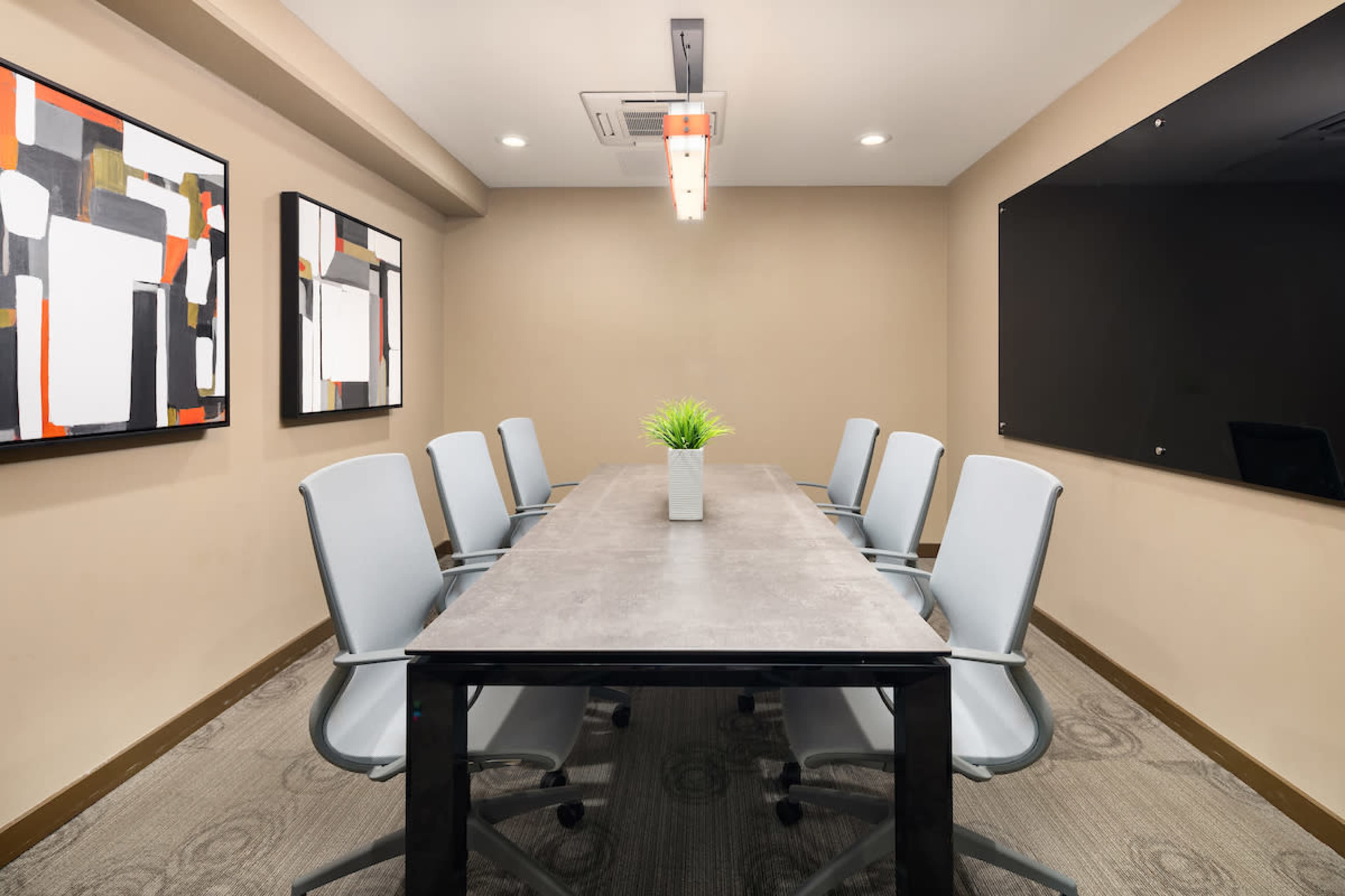 A modern conference room features a long table surrounded by six gray chairs, with artwork on the walls and a small plant centerpiece.