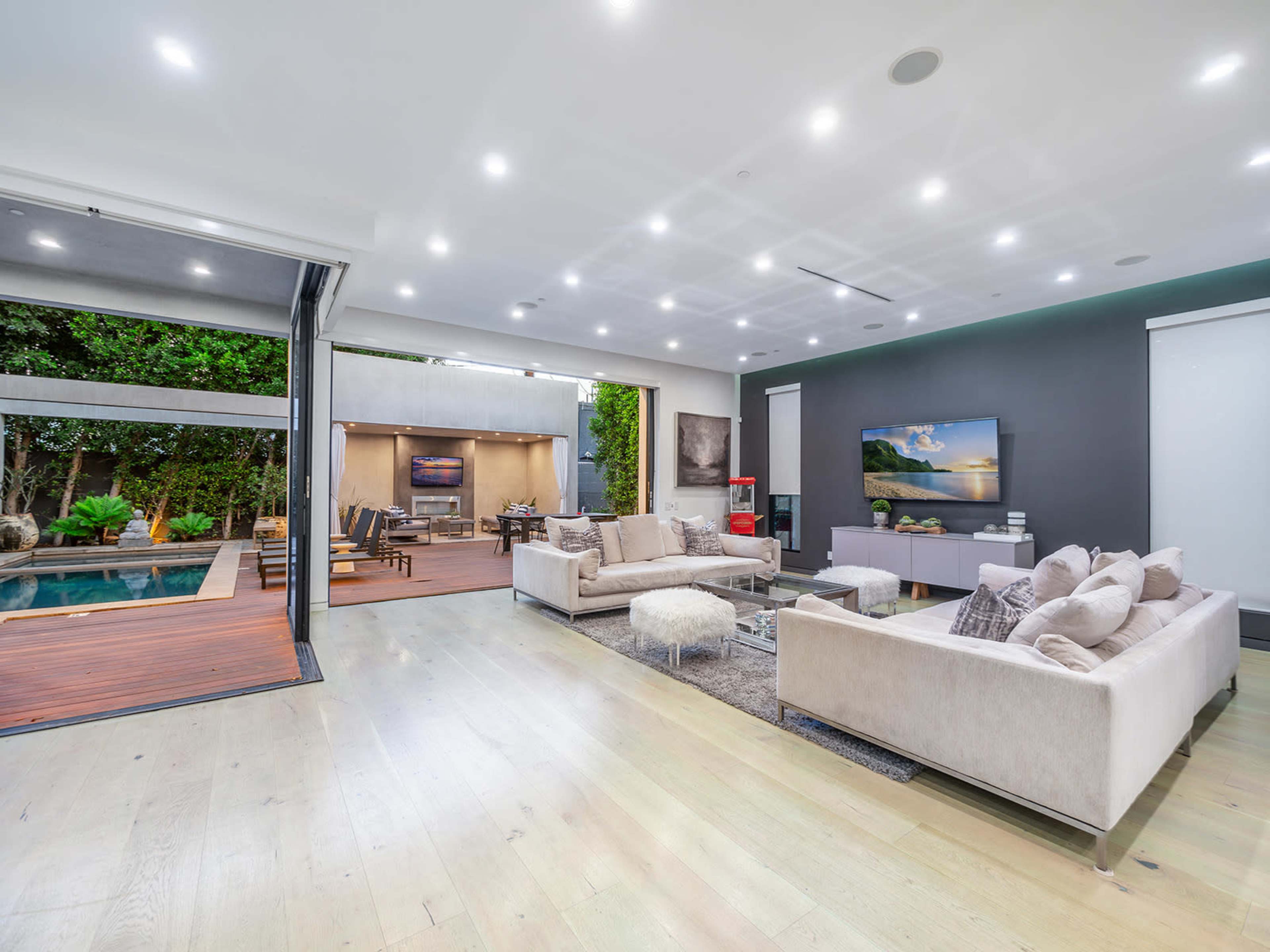 A modern living room that opens up to an outdoor space with a pool, featuring light-colored furniture and a large flat-screen TV.