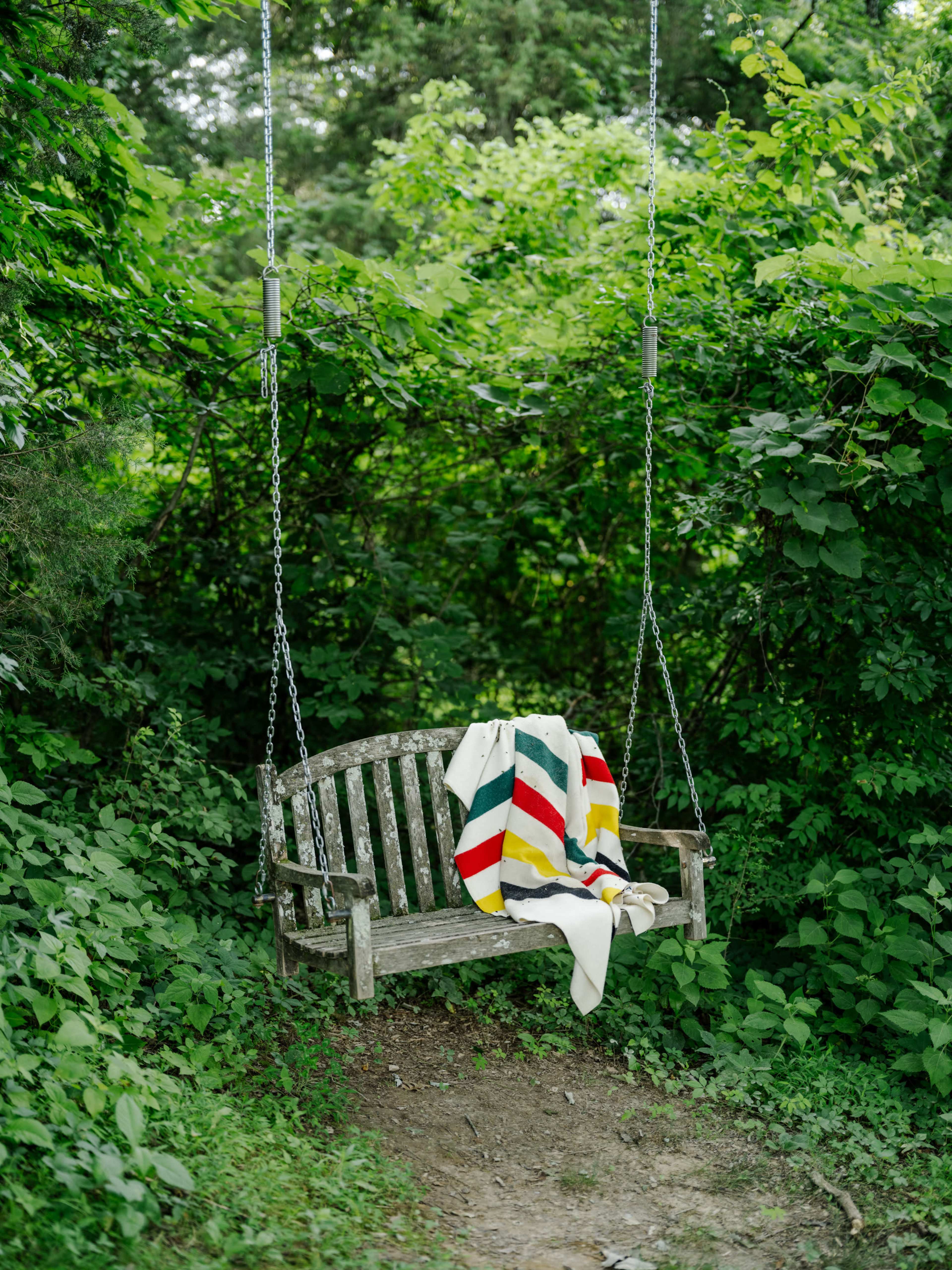 A wooden swing bench is surrounded by lush green foliage and draped with a colorful striped blanket.