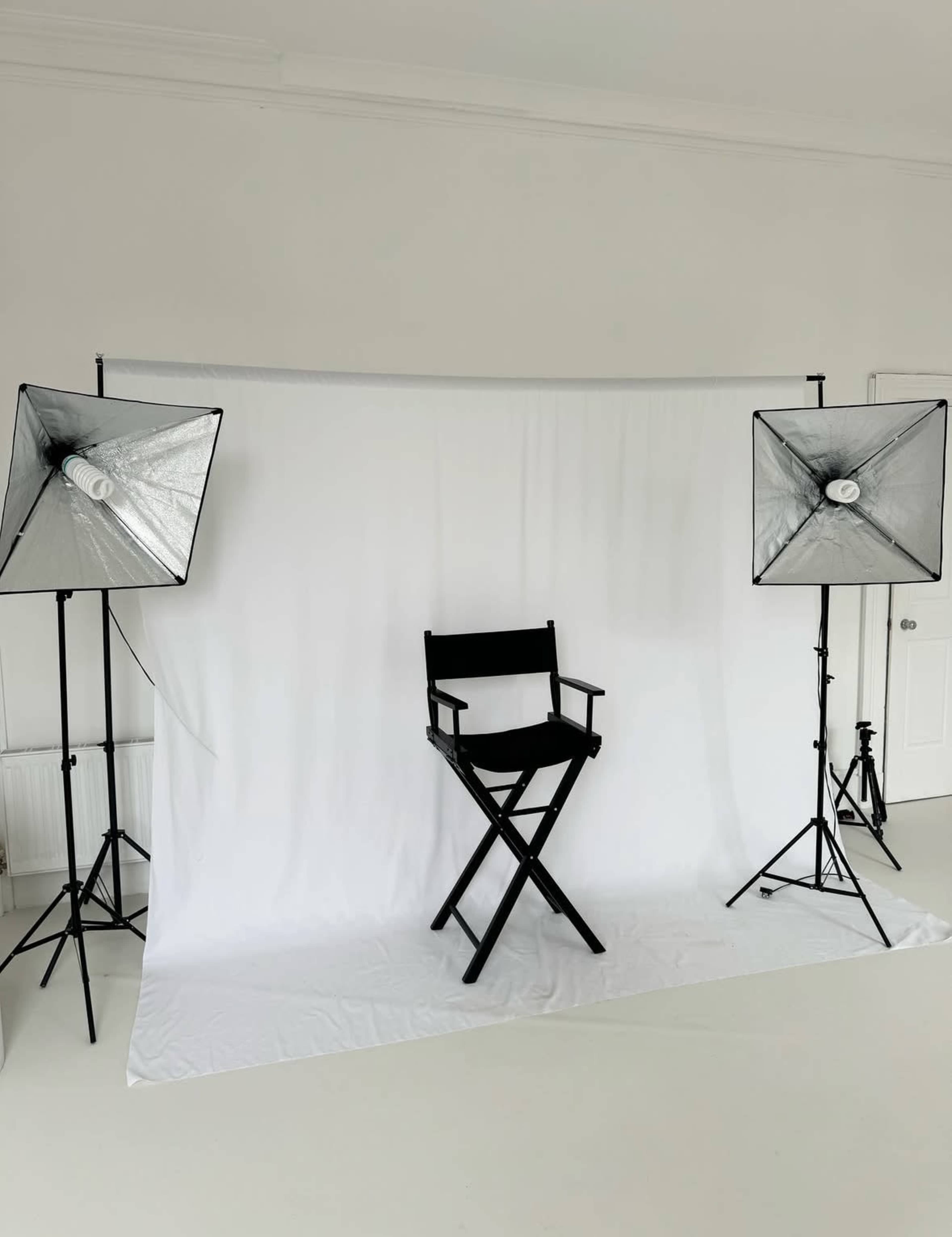 The image shows a photography studio setup with a white backdrop, a black director's chair in the center, and two light softboxes on either side.