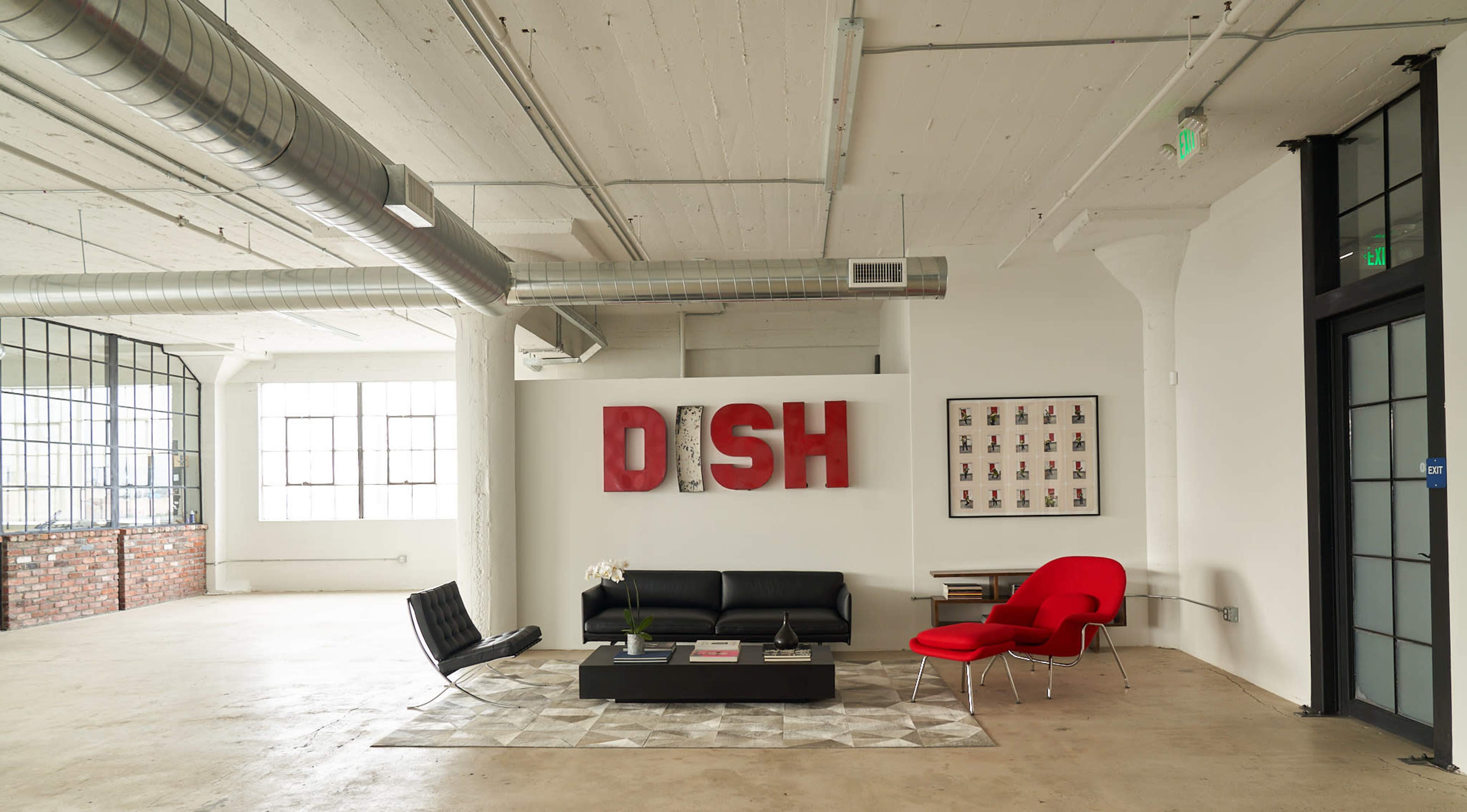A spacious, minimalist interior featuring a black leather sofa and a red chair, with large windows and a wall displaying the word "DISH" in bold red letters.