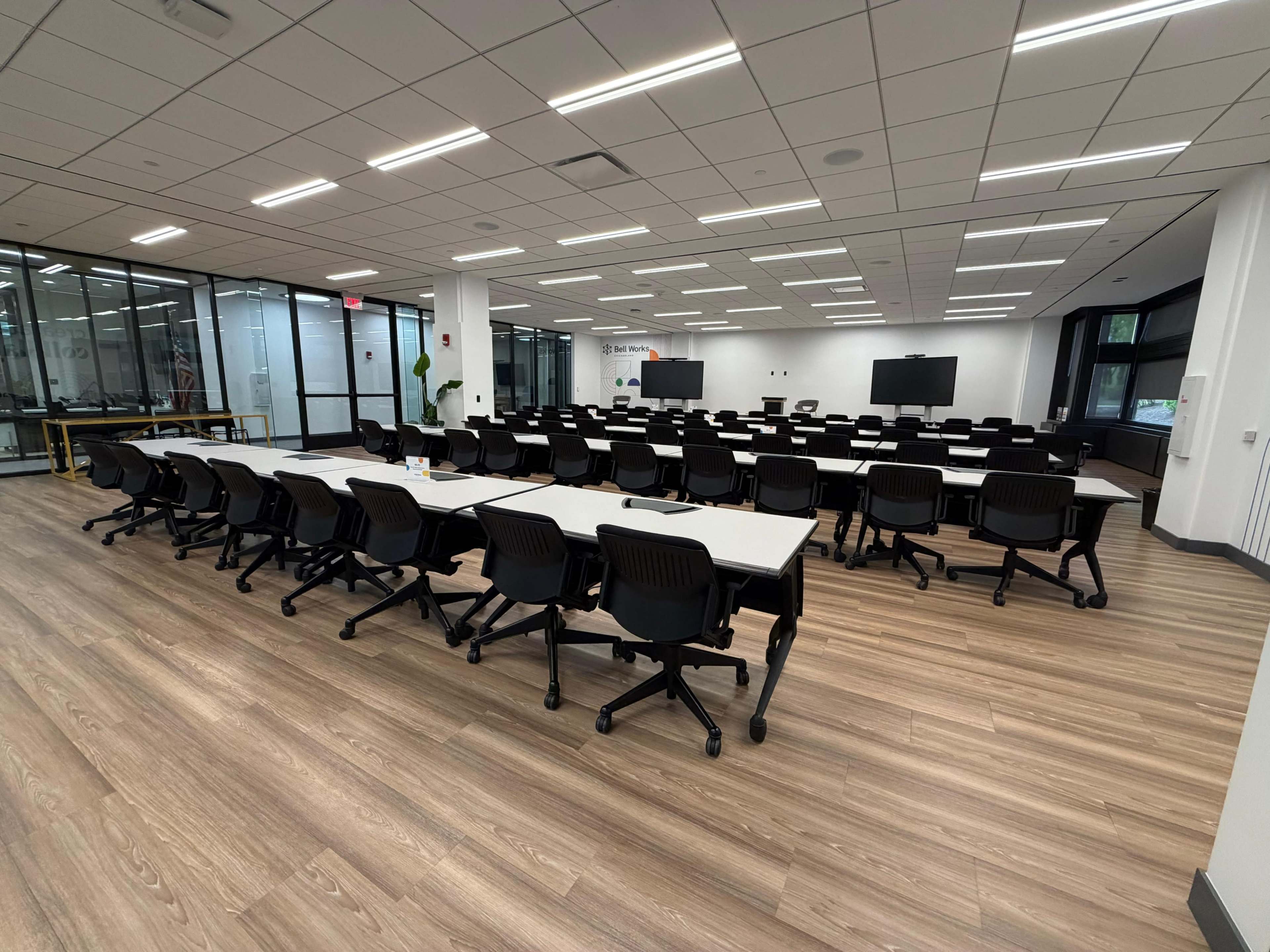 The image shows a modern classroom setup with rows of tables and chairs arranged on a wooden floor, featuring large windows and white walls.
