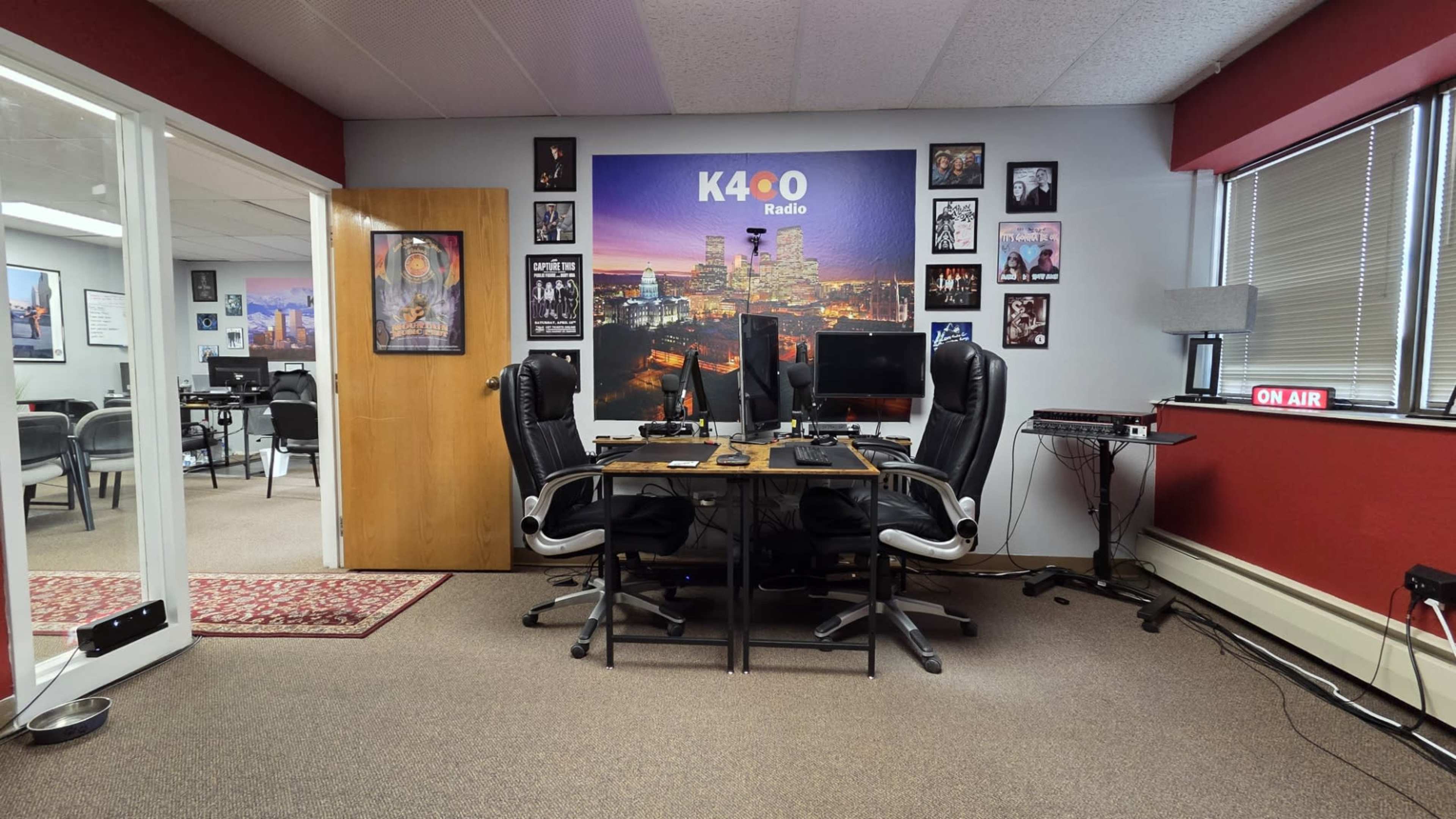 The image shows an office space with two desk chairs facing two computer monitors, surrounded by various photos and a "ON AIR" sign.