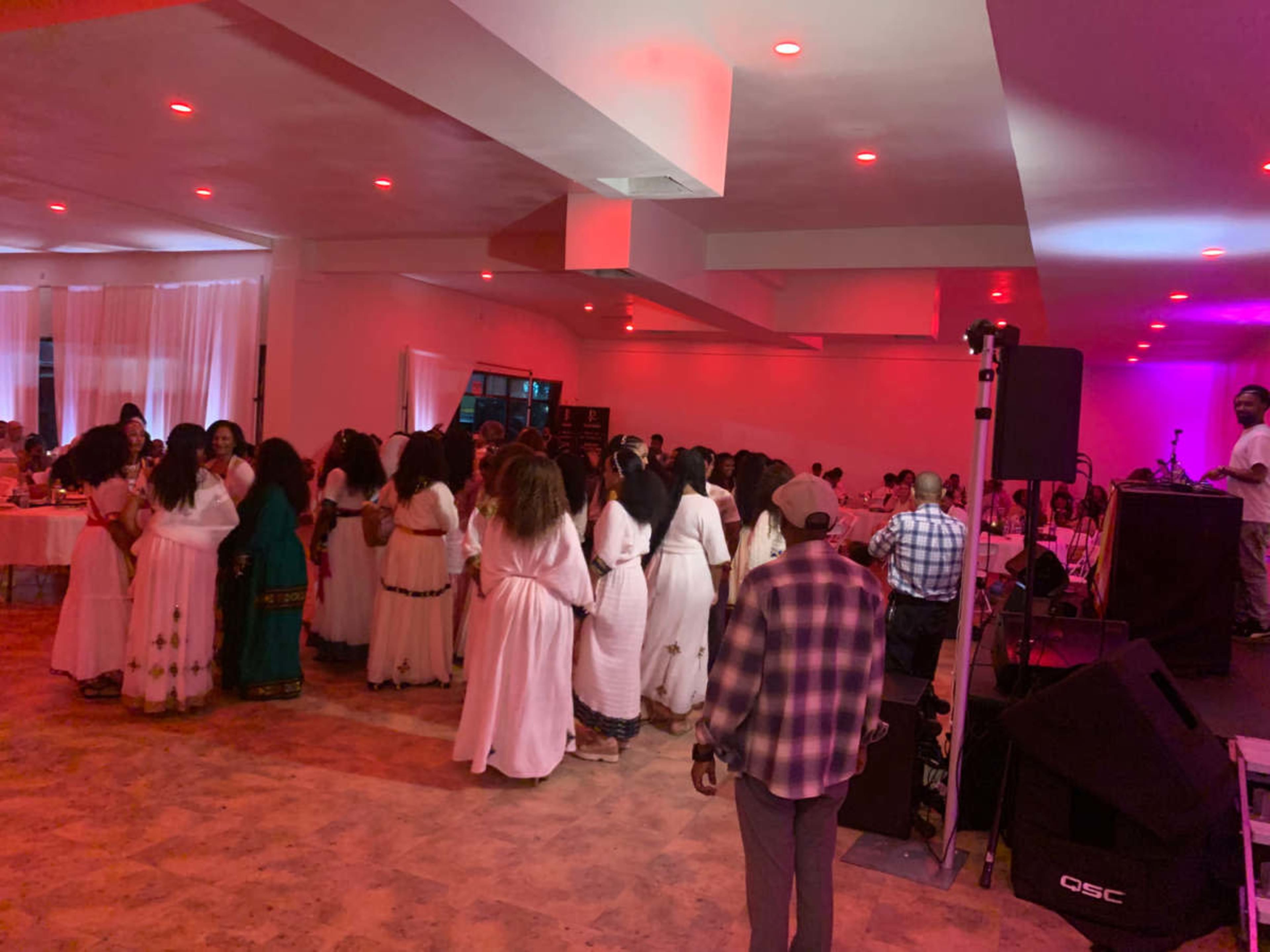A group of people wearing white dresses is dancing in a brightly lit room with a DJ setup in the background.