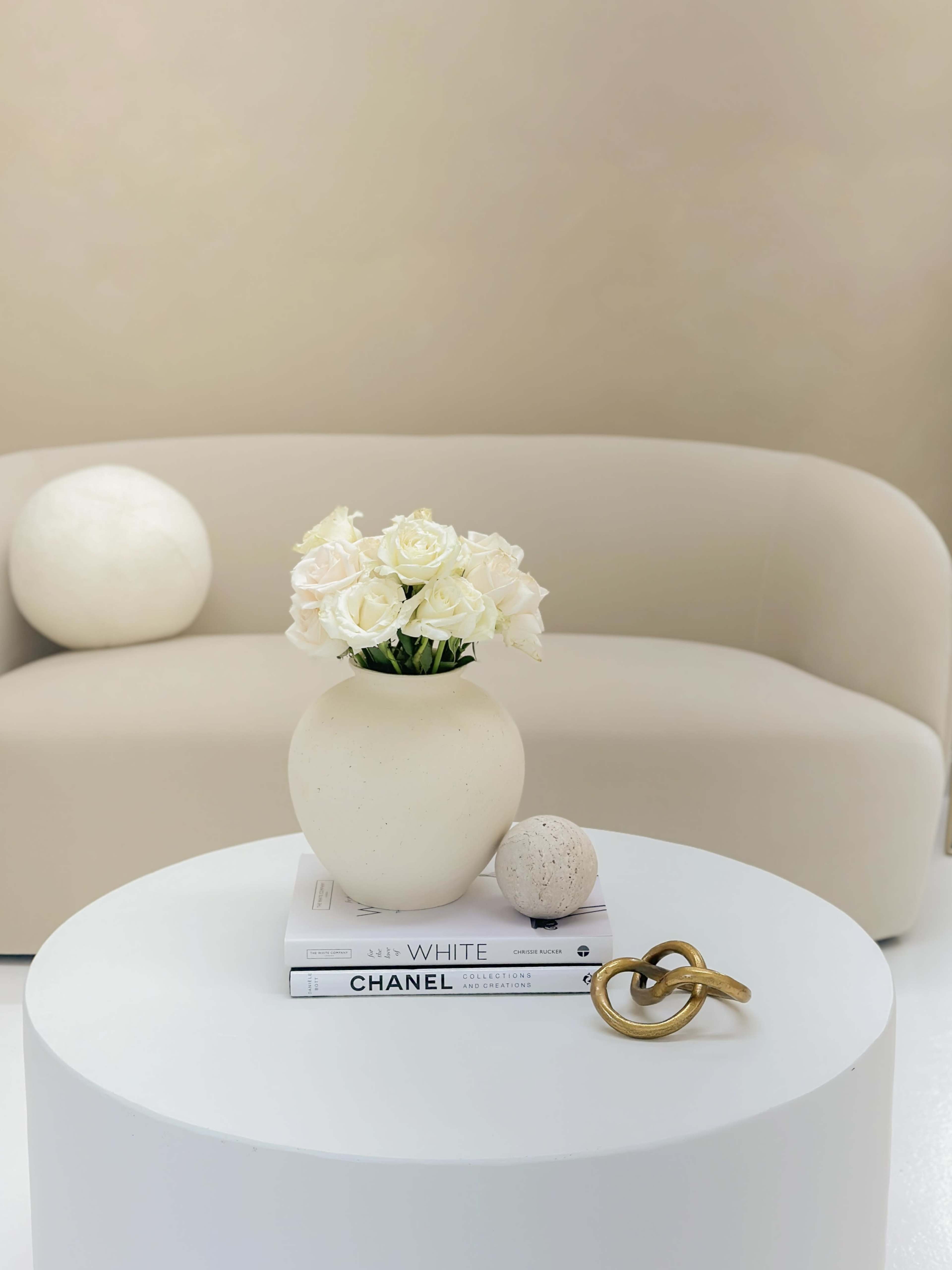 A white vase filled with white roses sits on a stack of books atop a round white table beside a curved beige sofa.