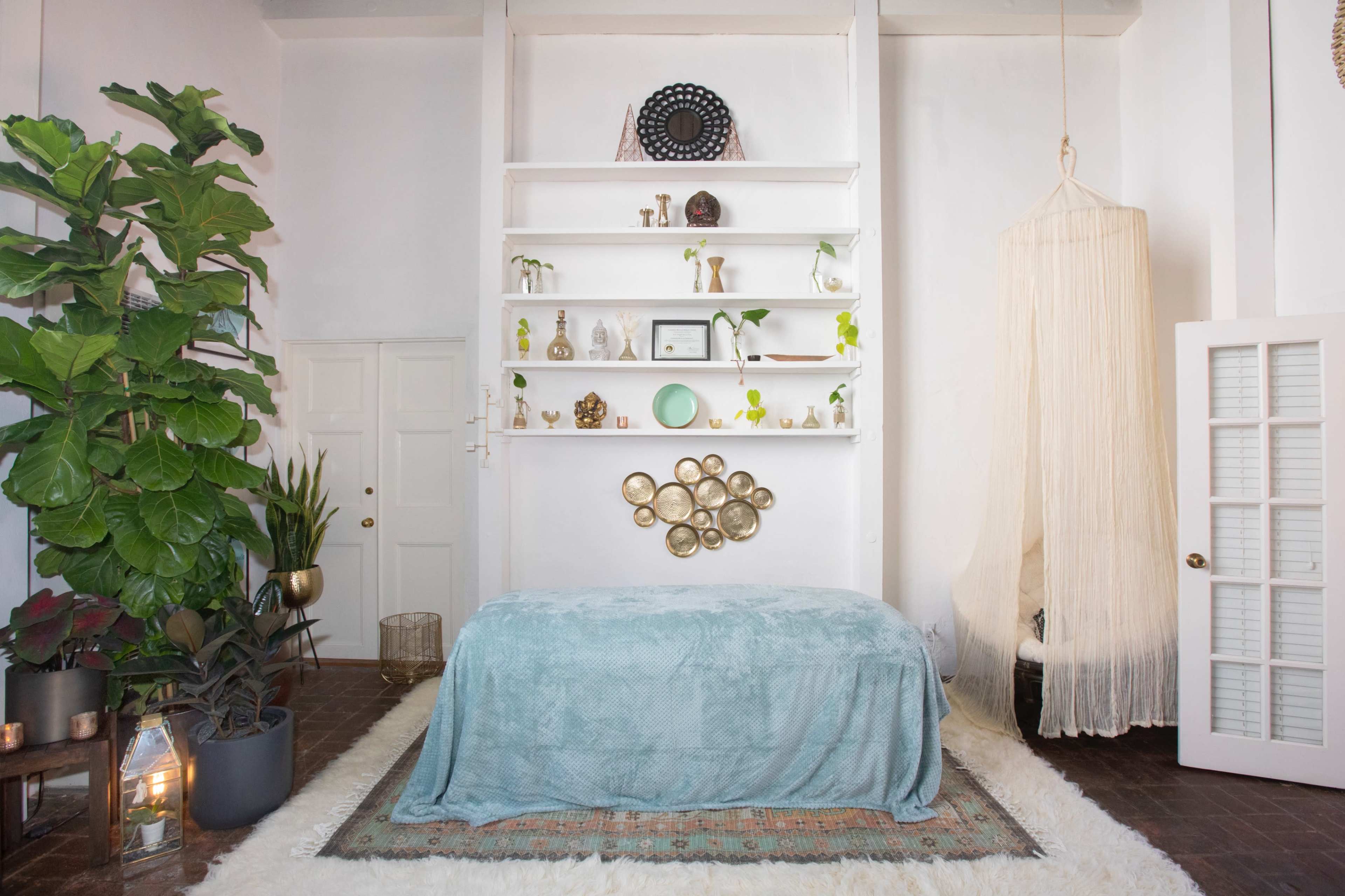 A serene room with a bed covered in a light blue blanket, surrounded by plants, decorative shelves, and a hanging draped fabric.