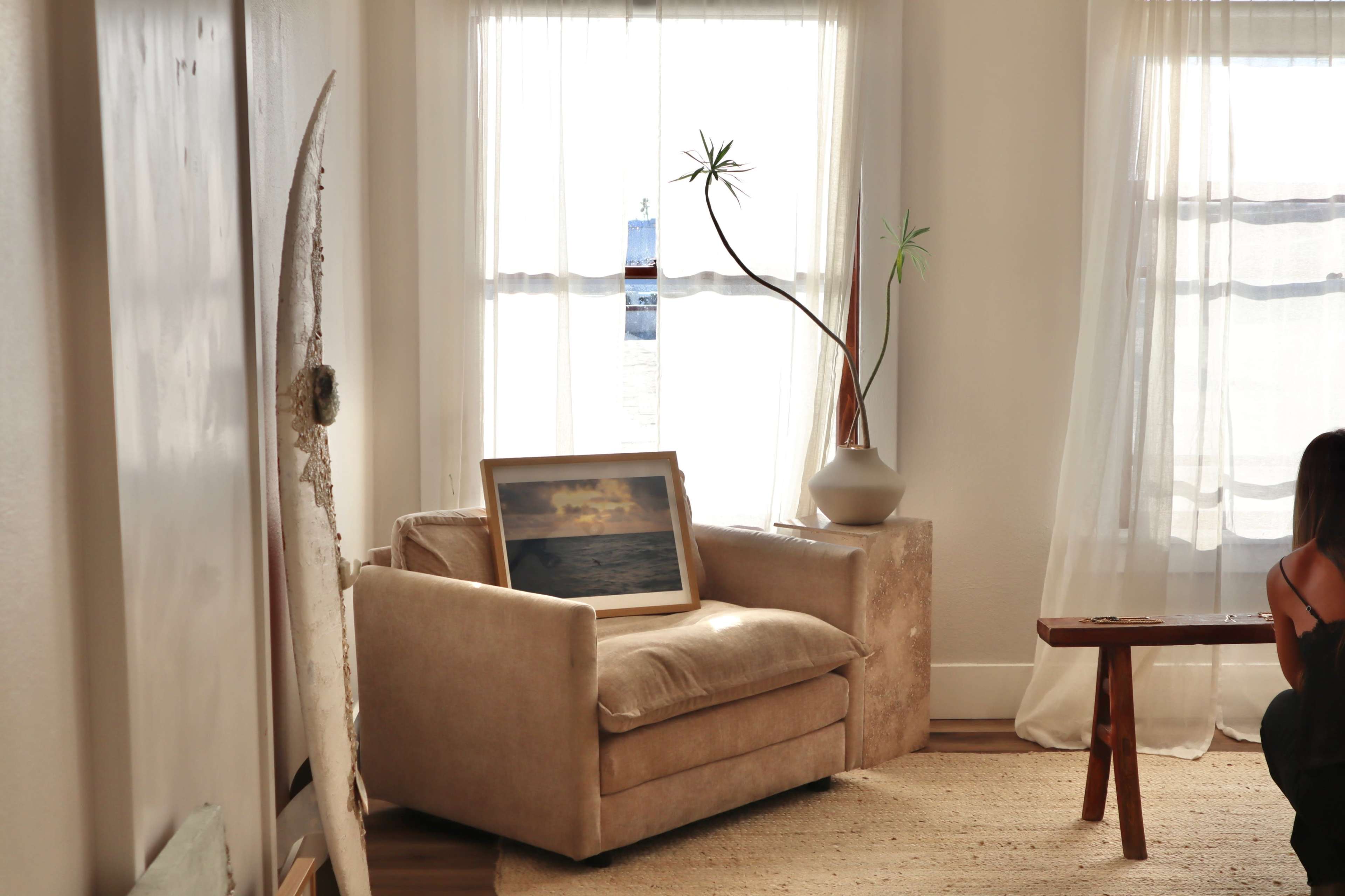 A beige armchair sits by a window, with a painting propped against it and a potted plant on a nearby table.