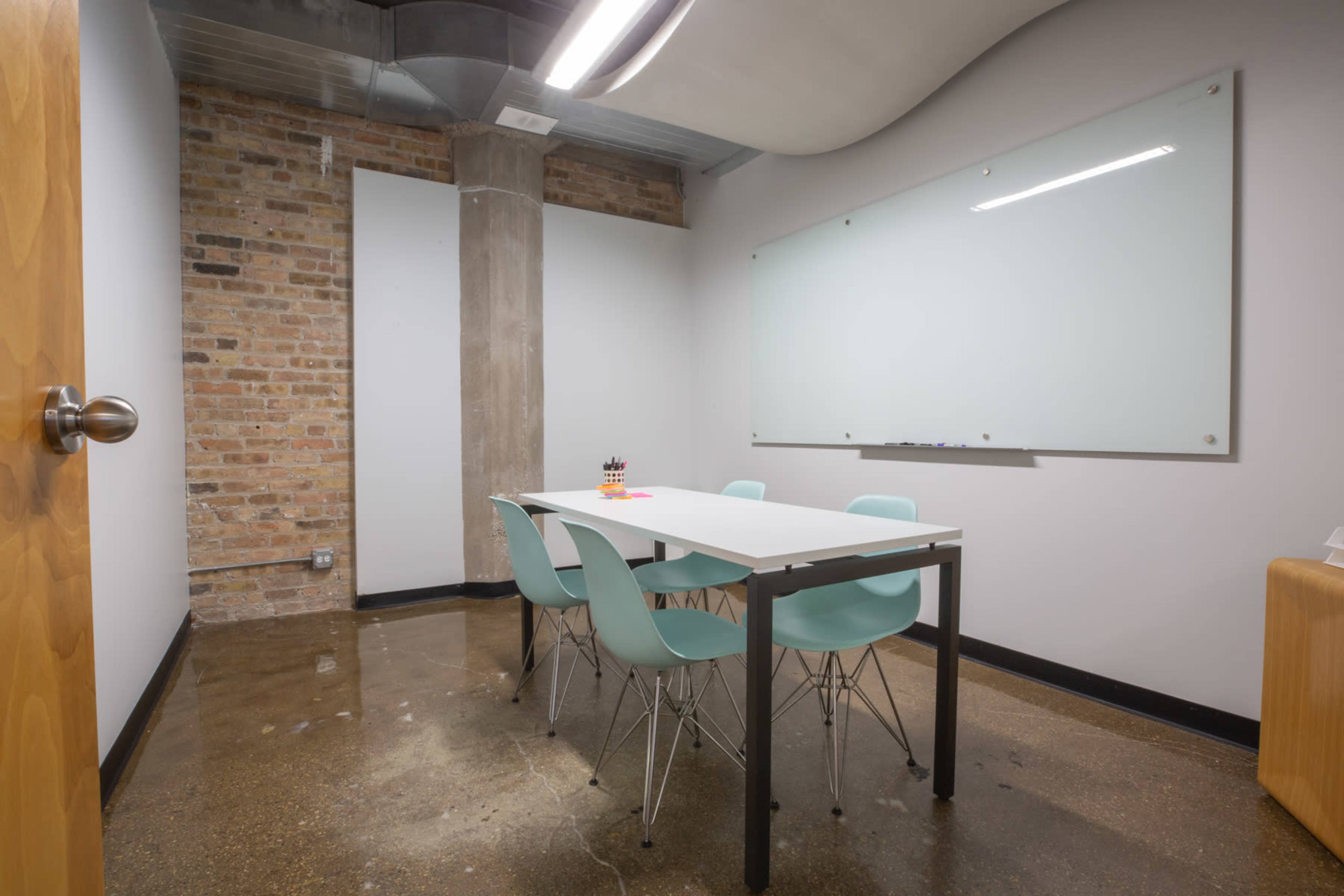 A small conference room features a white table surrounded by light blue chairs and a large whiteboard on the wall.
