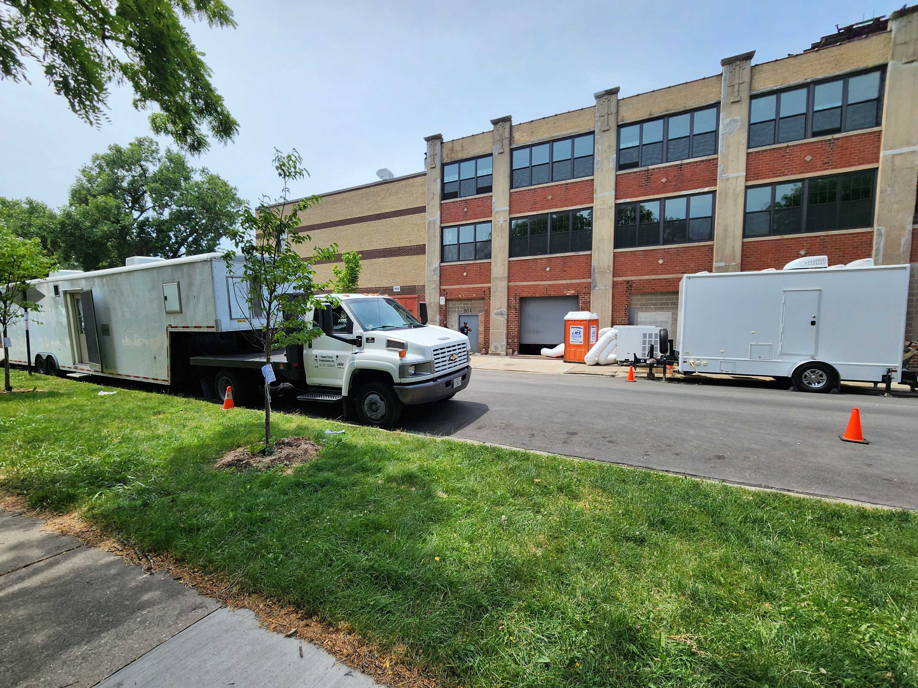 A white truck is parked on a street in front of a brick building, accompanied by several trailers and portable restrooms.
