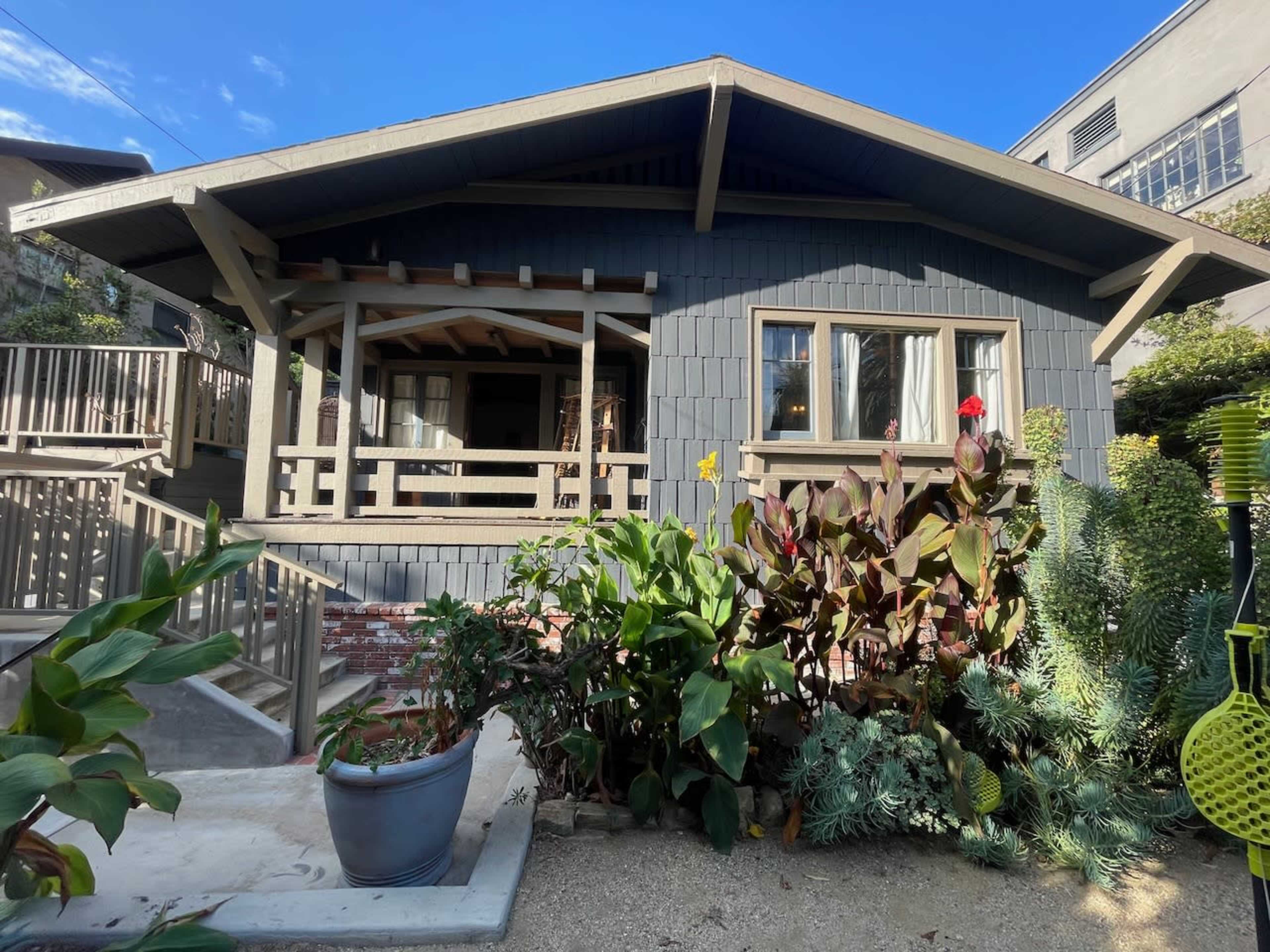 The image shows a single-story craftsman-style house with a gray exterior, surrounded by decorative plants and a gravel pathway.