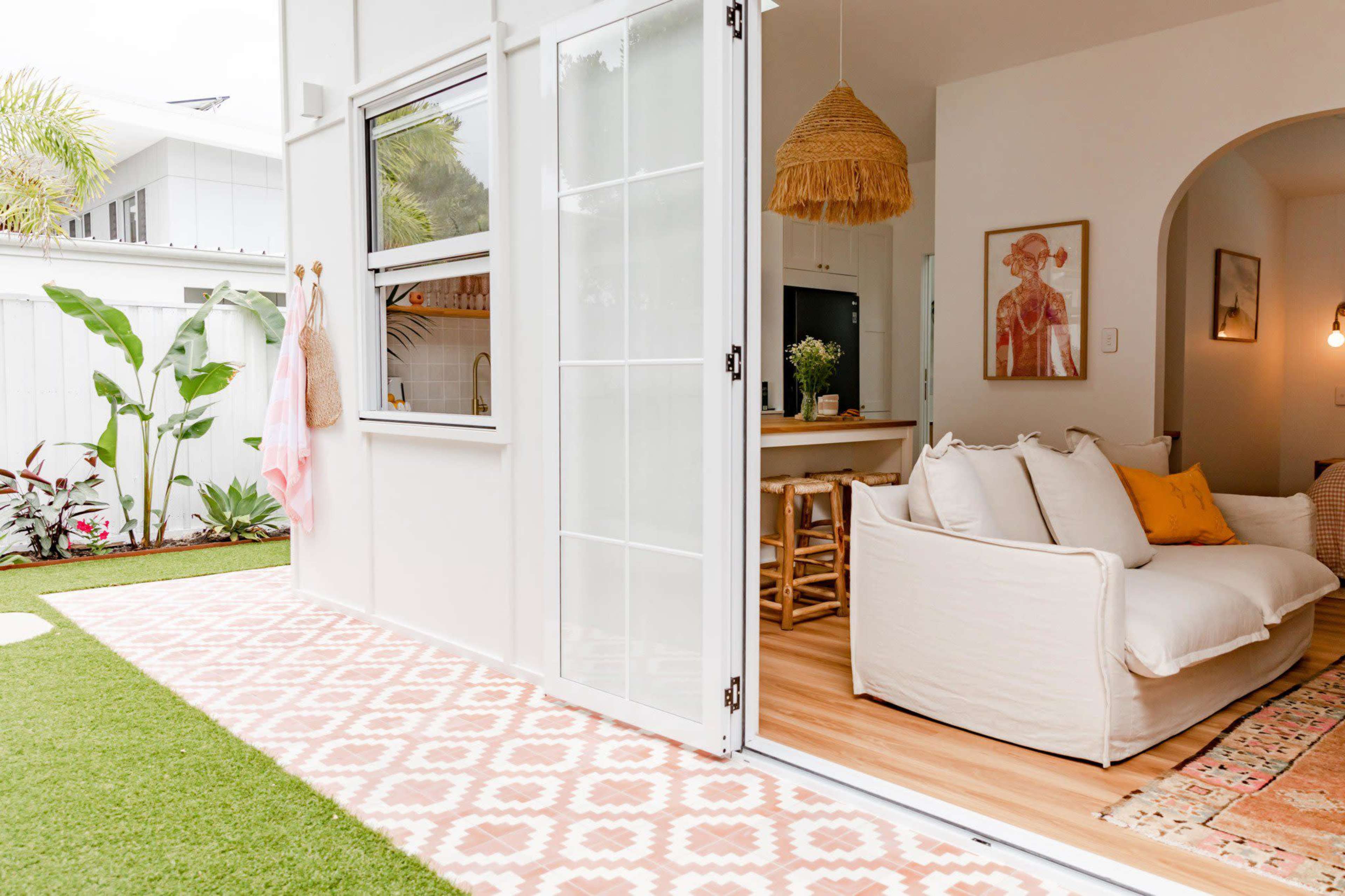 A bright living space features an open door leading to a patio with laid tile and a green lawn, showcasing a cozy couch and a kitchen area in the background.