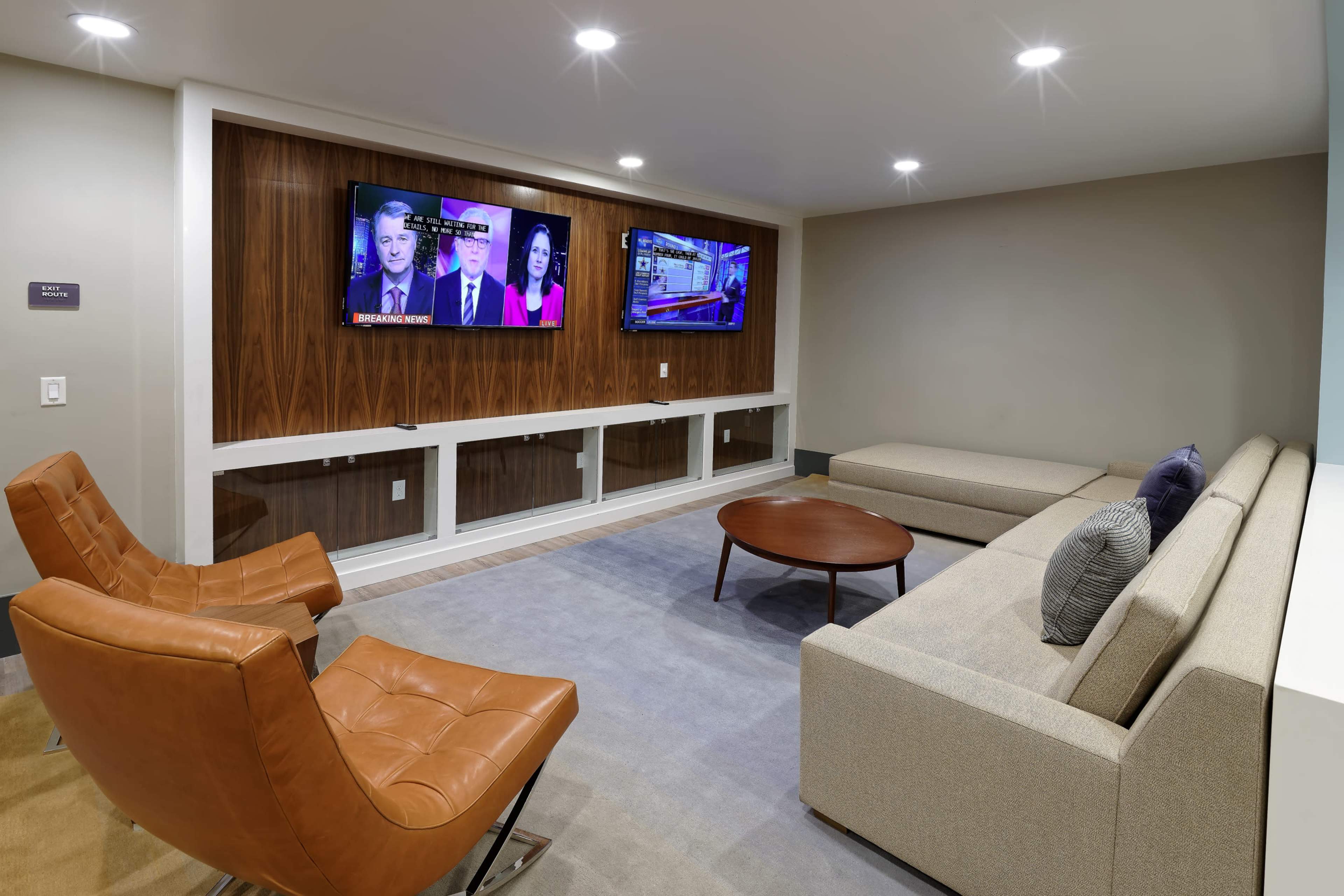 A modern lounge area with two television screens mounted on a wooden wall, seating arrangements comprised of two orange chairs and a gray sofa, and a round coffee table in the center.