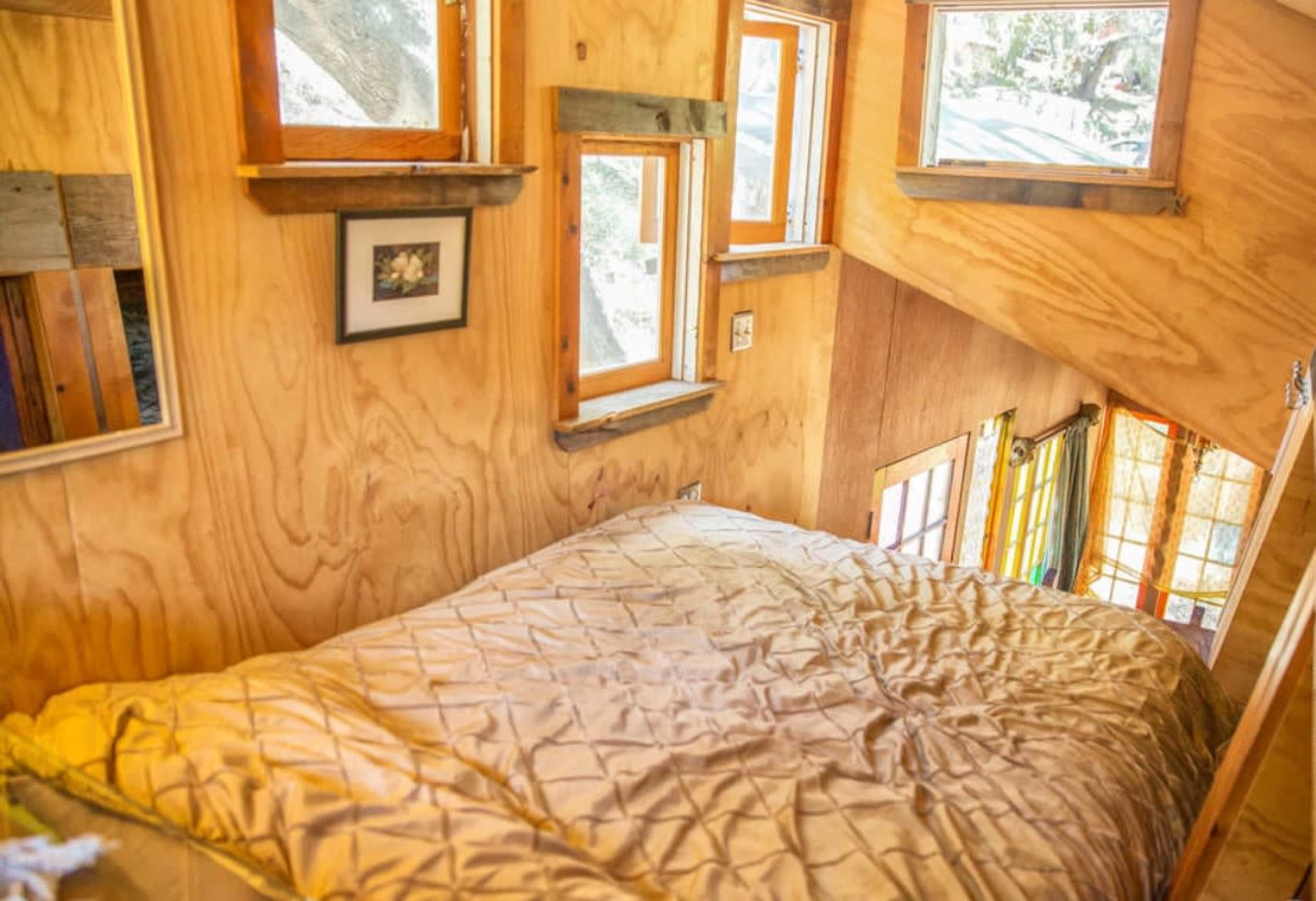 A small, wooden interior of a lofted bedroom with multiple windows, a bed covered in a light quilt, and wooden shelves holding framed pictures.