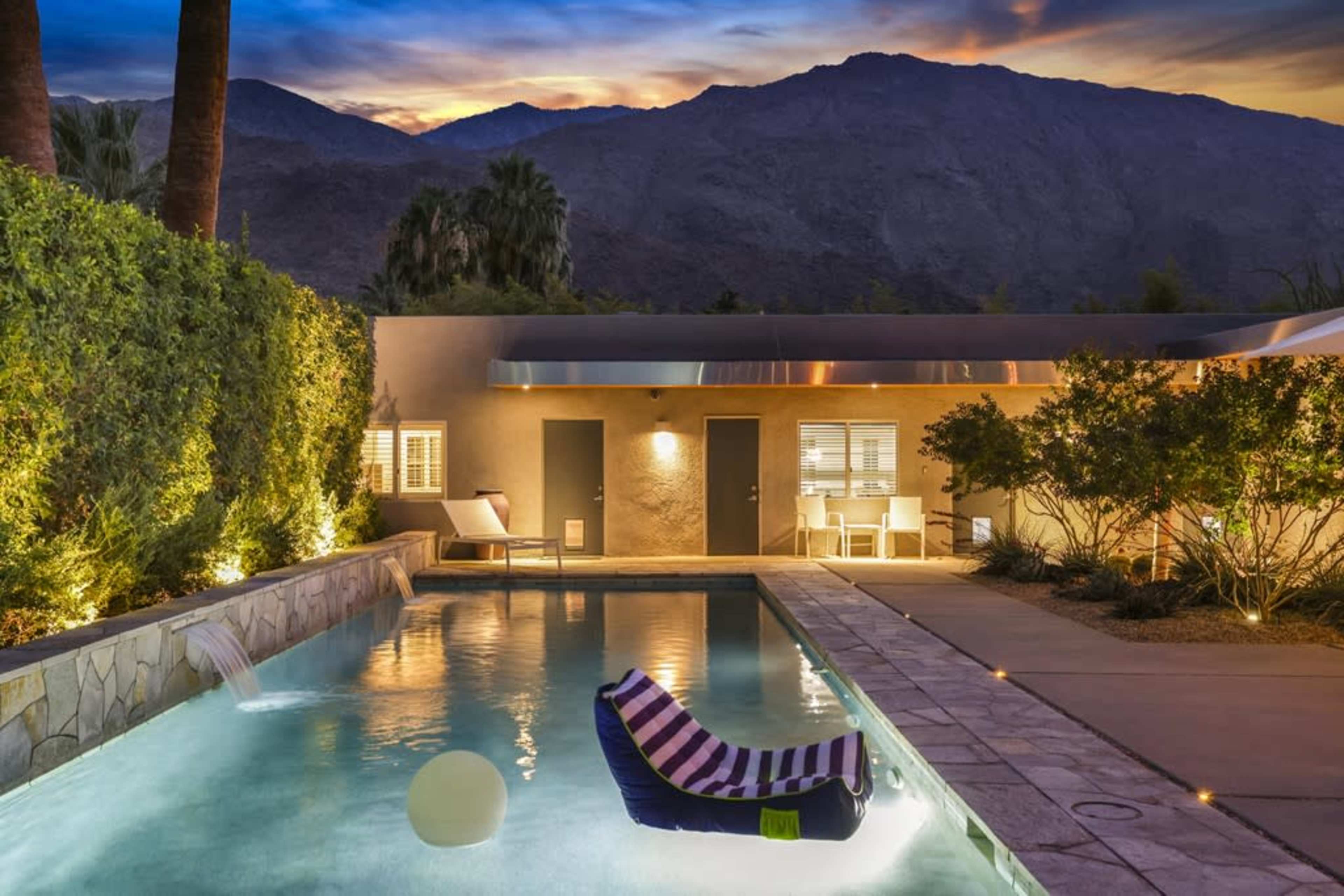 The image shows a tranquil pool area with a lounge chair and a floating inflatable in front of a mountain backdrop at sunset.