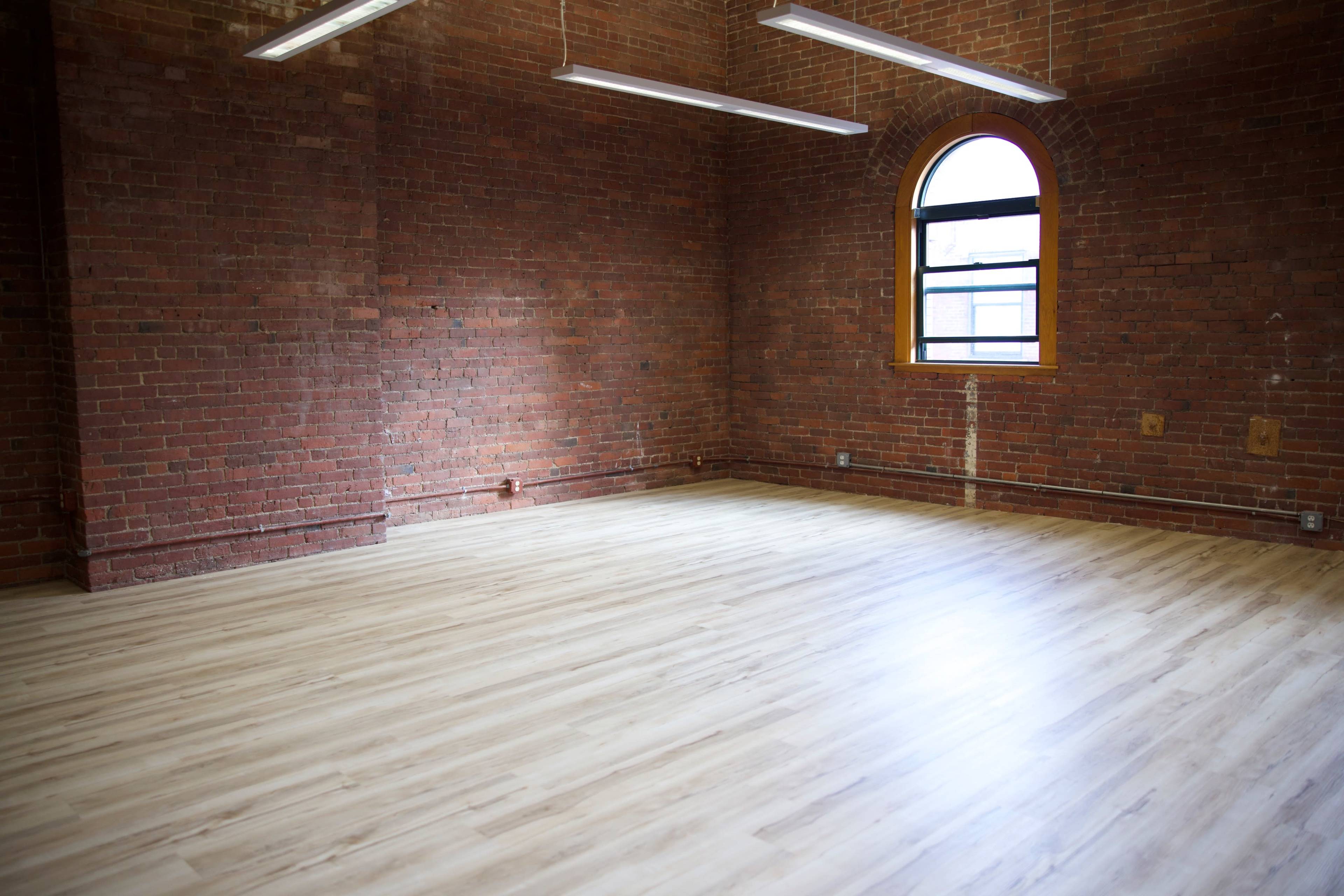 An empty room with exposed brick walls and a large window letting in natural light, featuring a wooden floor.