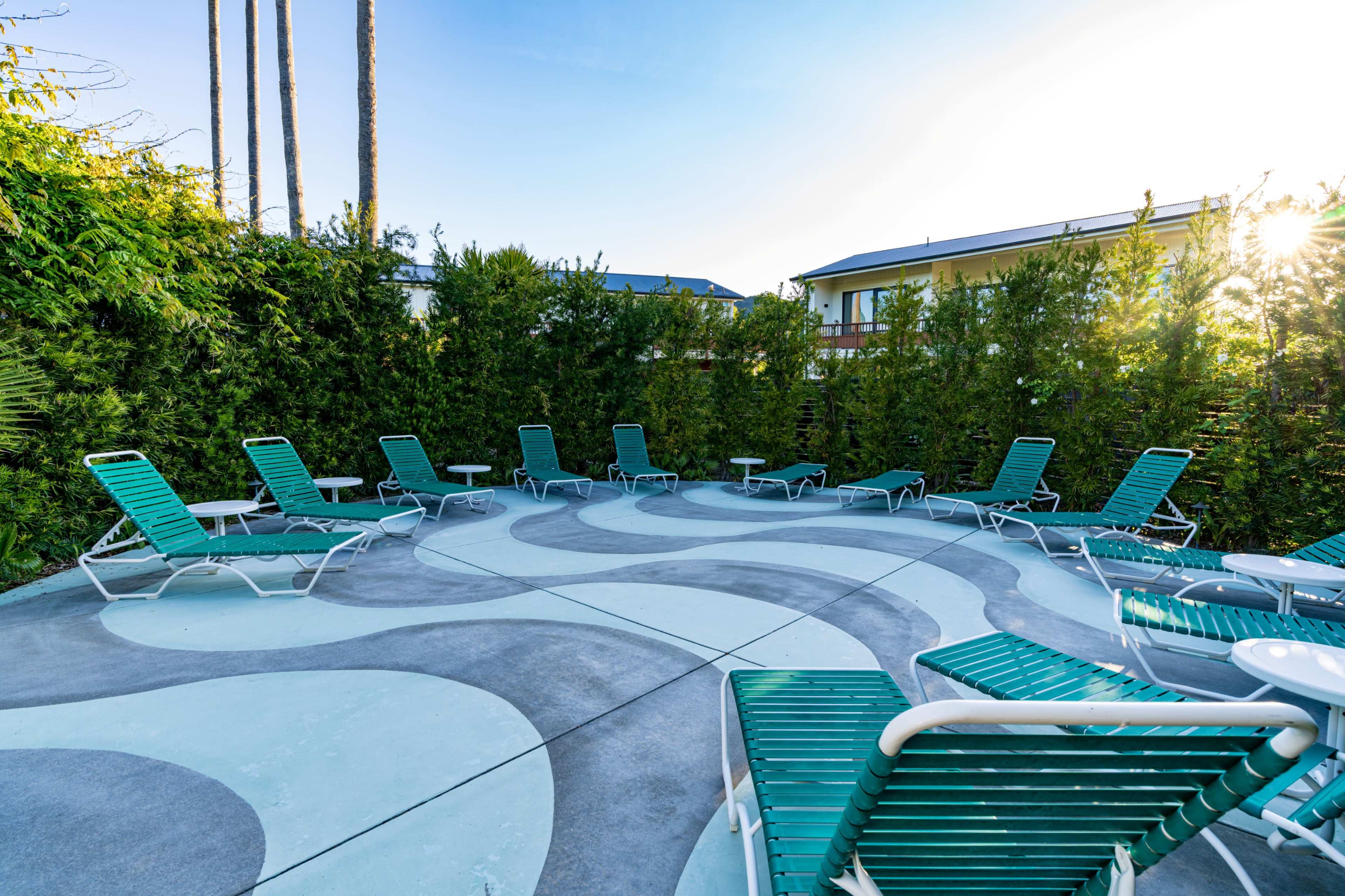 The image shows a circular seating area with several green lounge chairs arranged on a patterned concrete surface, surrounded by tall greenery and a building in the background.