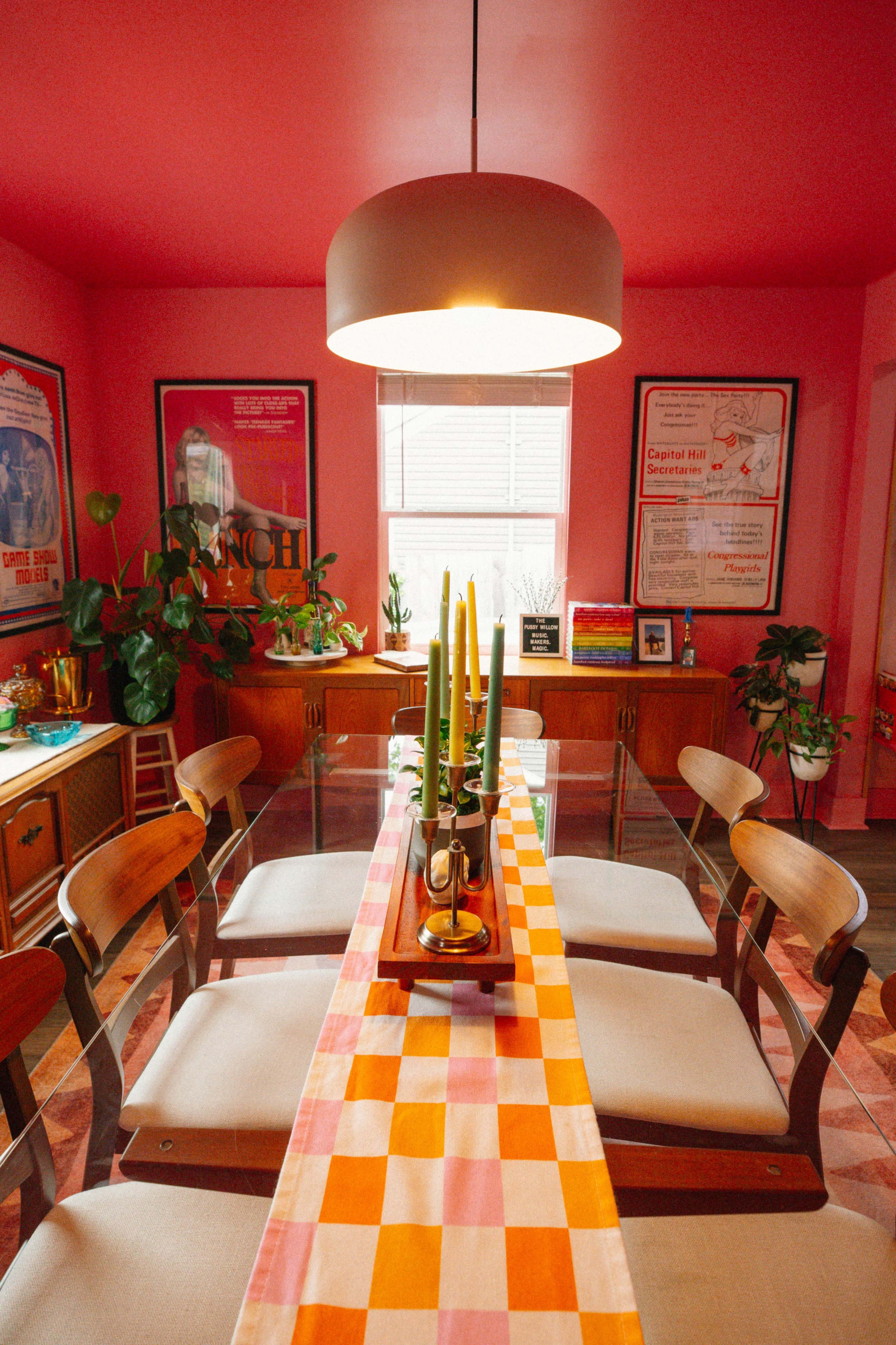 A dining room features a glass table with a colorful checkered table runner, surrounded by wooden chairs and illuminated by a pendant light, with plants and framed posters on the walls.